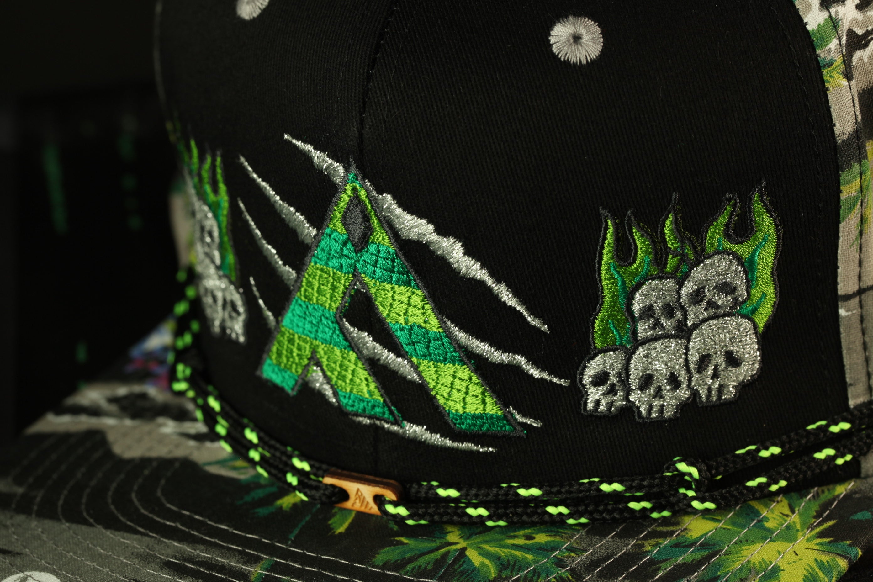 a close-up view of a black baseball cap with various embroidered designs and patterns, including a large green and black design featuring a skull and a flame-like element.