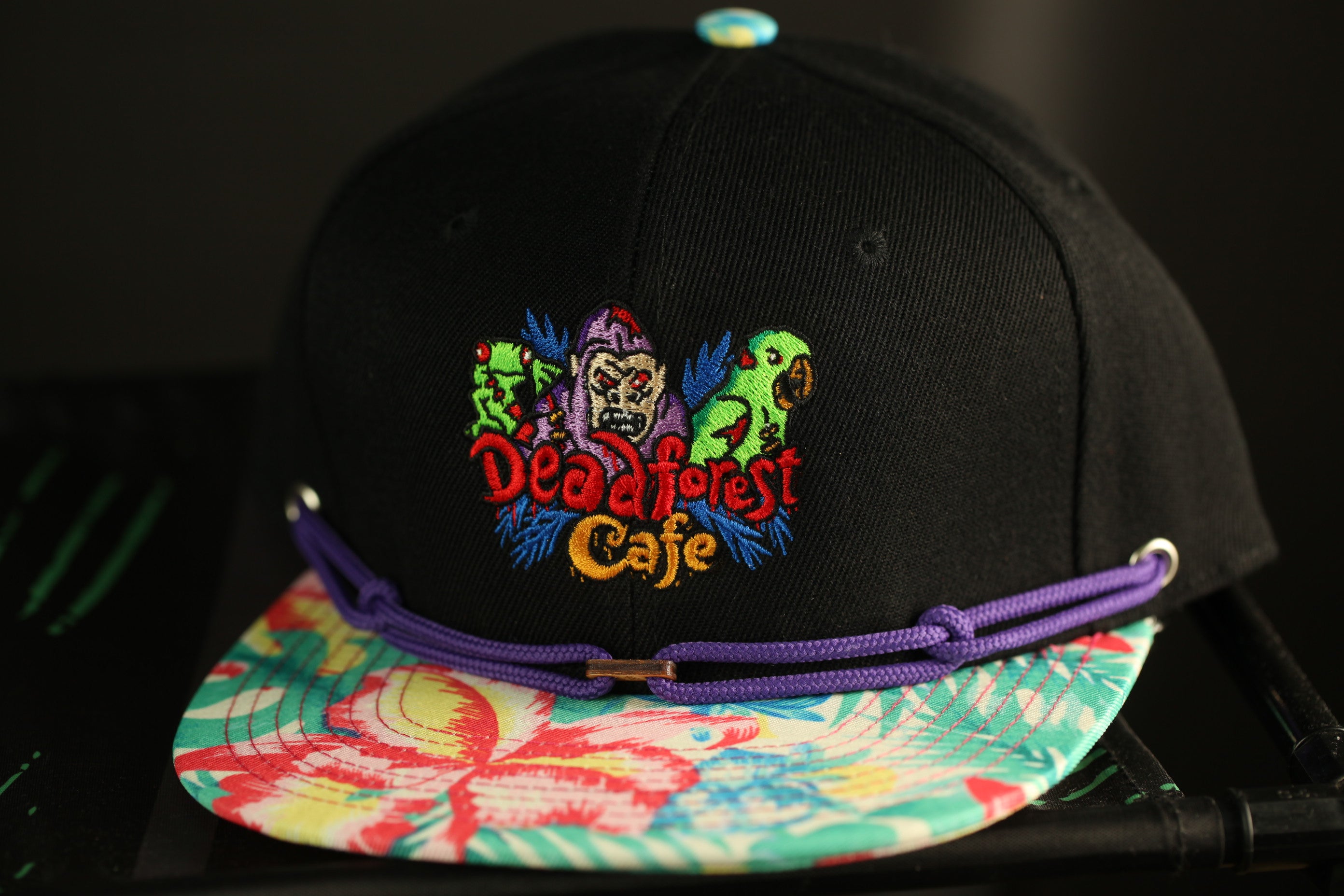 A black baseball cap with a colorful floral pattern and a purple band around it, featuring an embroidered logo for "Deadforest Cafe".