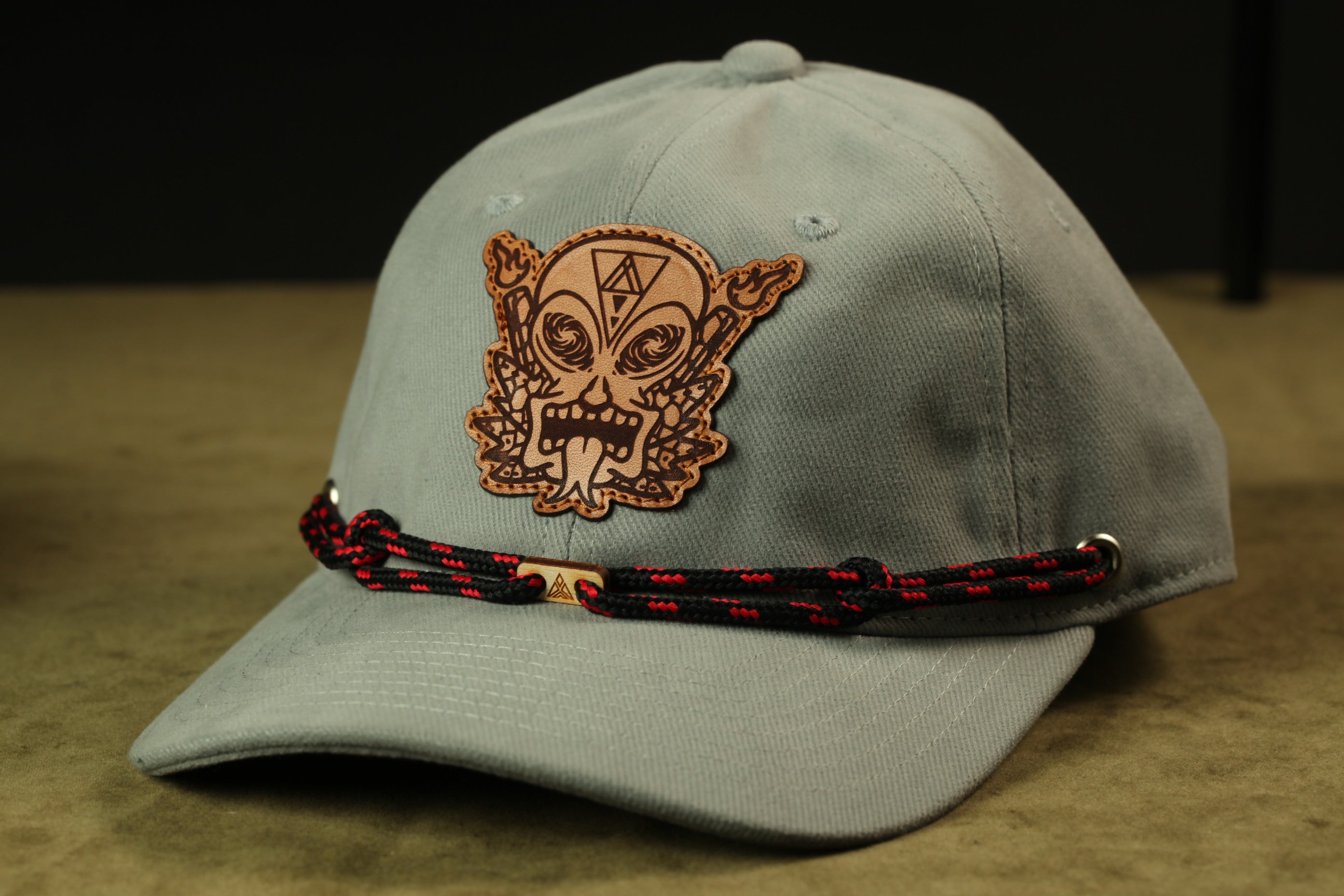 A gray baseball cap with a leather patch featuring a skull design and a red and black braided strap.