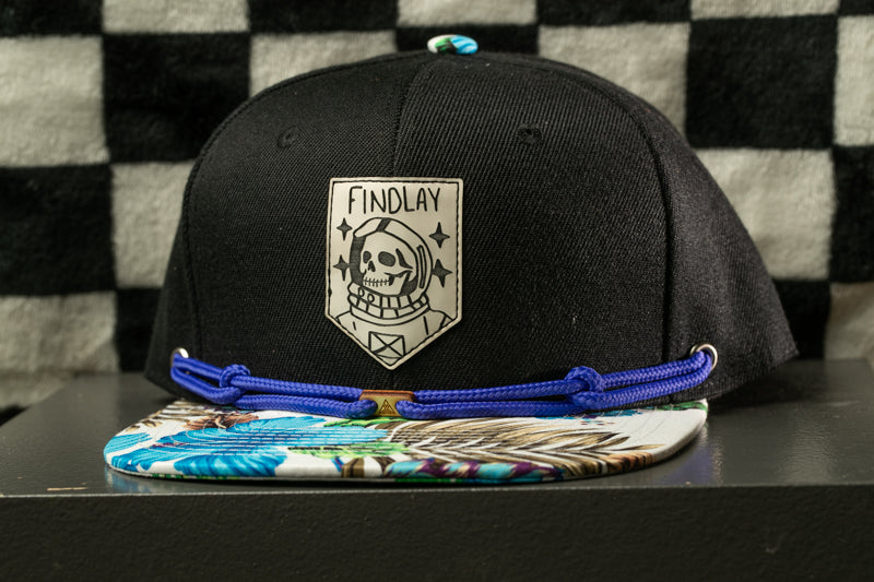 A black baseball cap with a white skull and crossbones design, and a blue string around the brim, is displayed against a black and white checkered background.