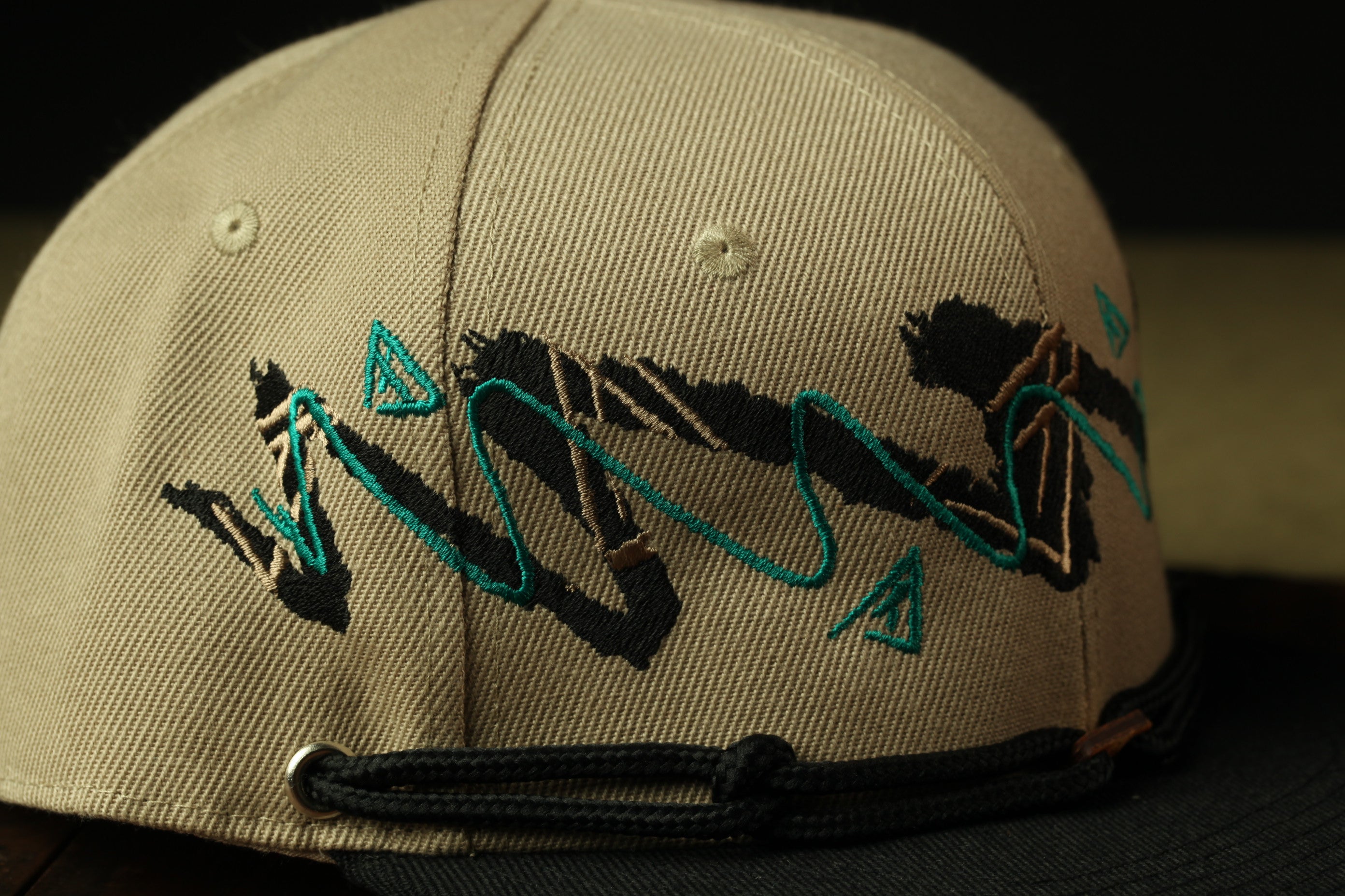 A close-up view of a beige baseball cap with a black band and a decorative embroidered design in turquoise and black on the front.