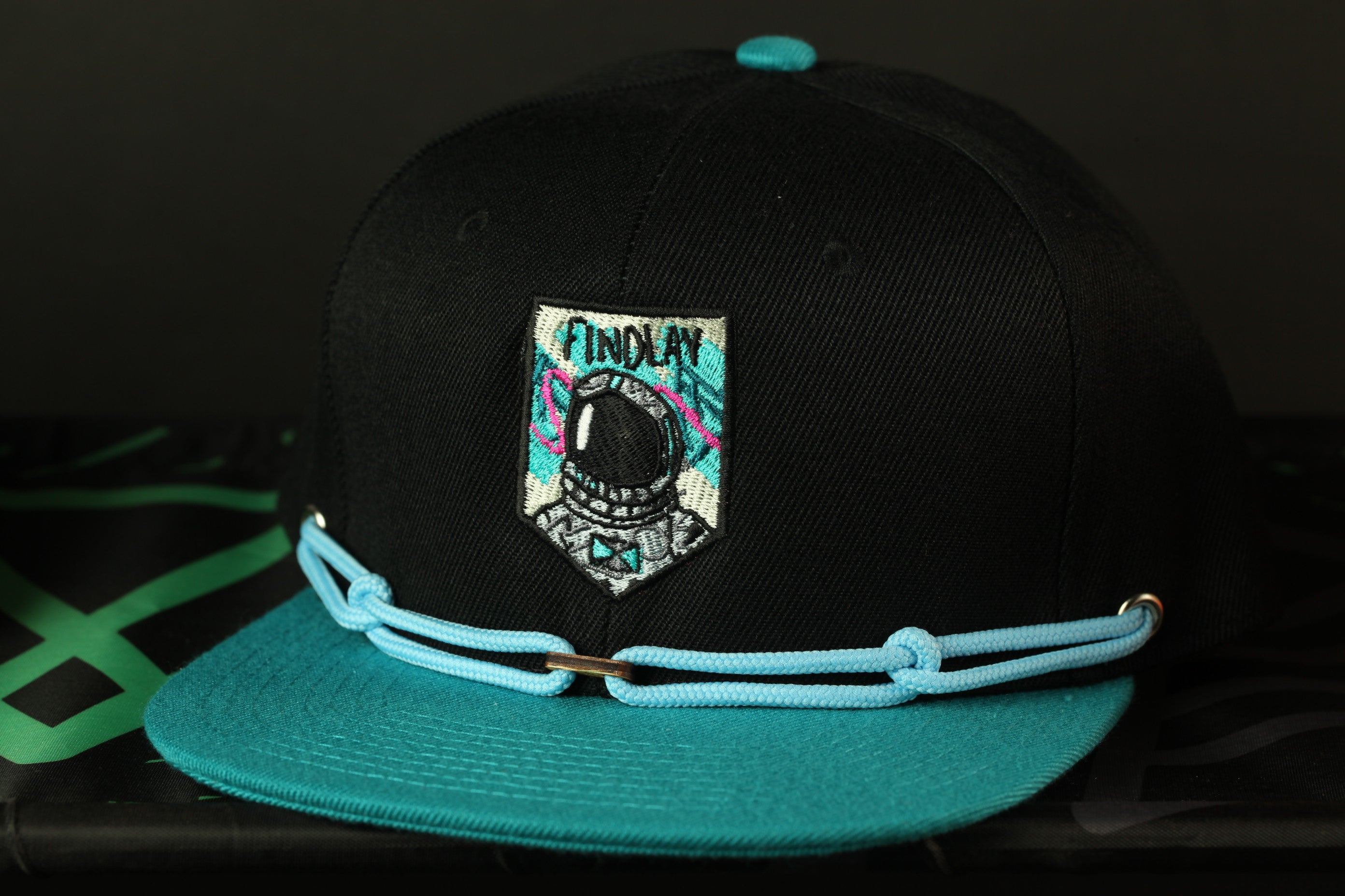 A black baseball cap with a teal brim and a patch featuring a space helmet design.