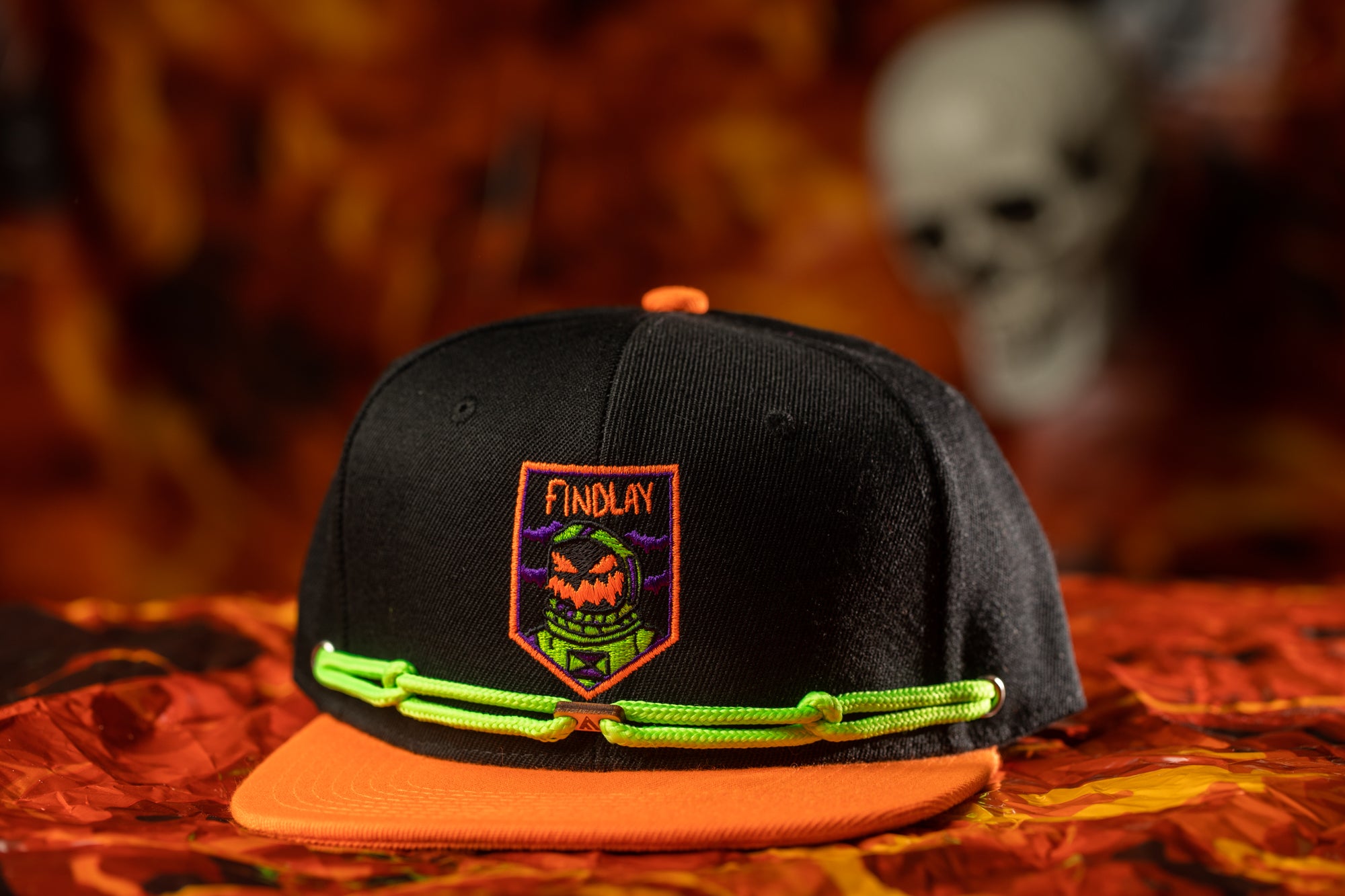 A black baseball cap with a green and orange band, featuring a patch with a skull and crossbones design, is displayed against a red and orange background.