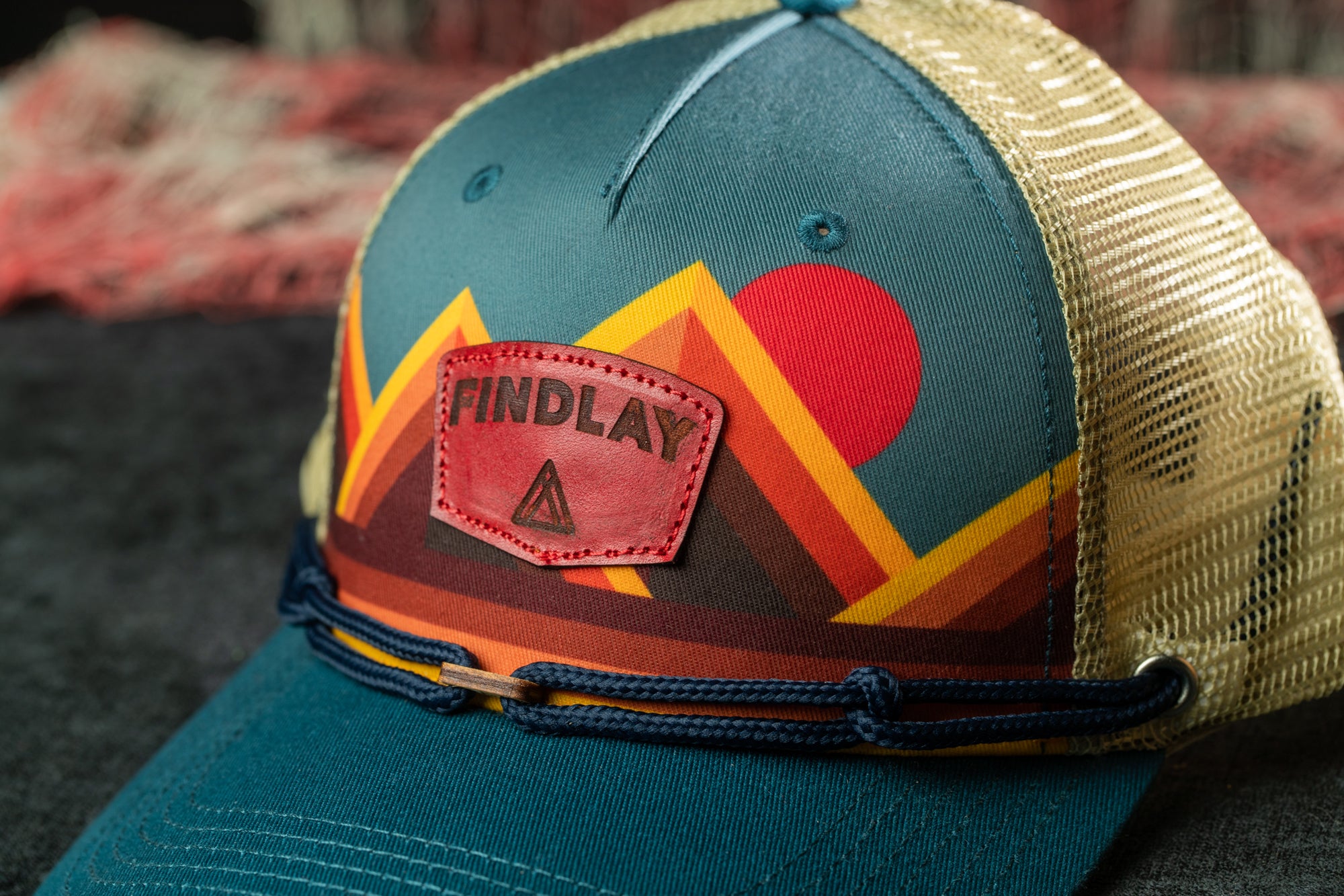 A close-up image of a colorful baseball cap with a mountain and sun motif, featuring a red triangle logo on the front.