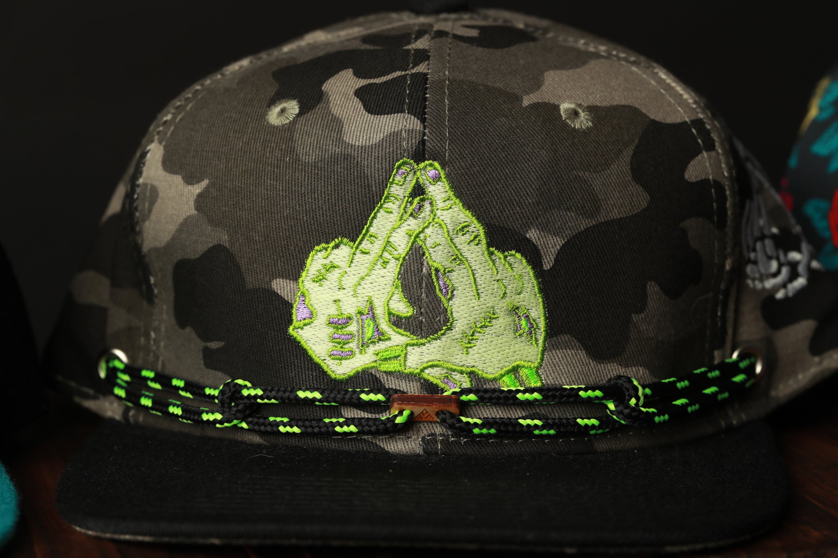 A camouflage baseball cap with a green and black braided strap and a unique embroidered design on the front.