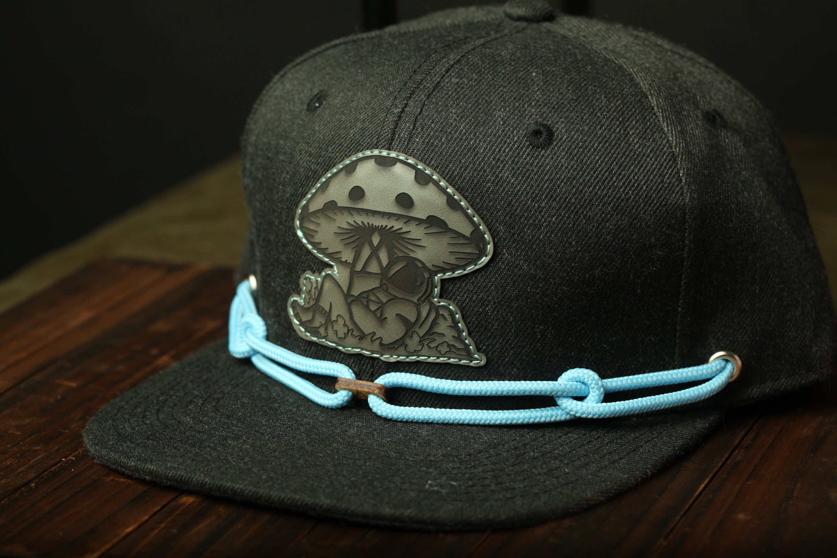 A black baseball cap with a mushroom patch and blue braided straps is displayed on a wooden surface.