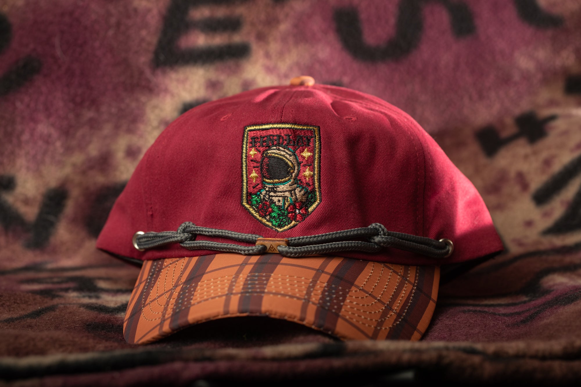 A red baseball cap with a plaid pattern and an embroidered patch featuring an astronaut is displayed against a purple and black background.