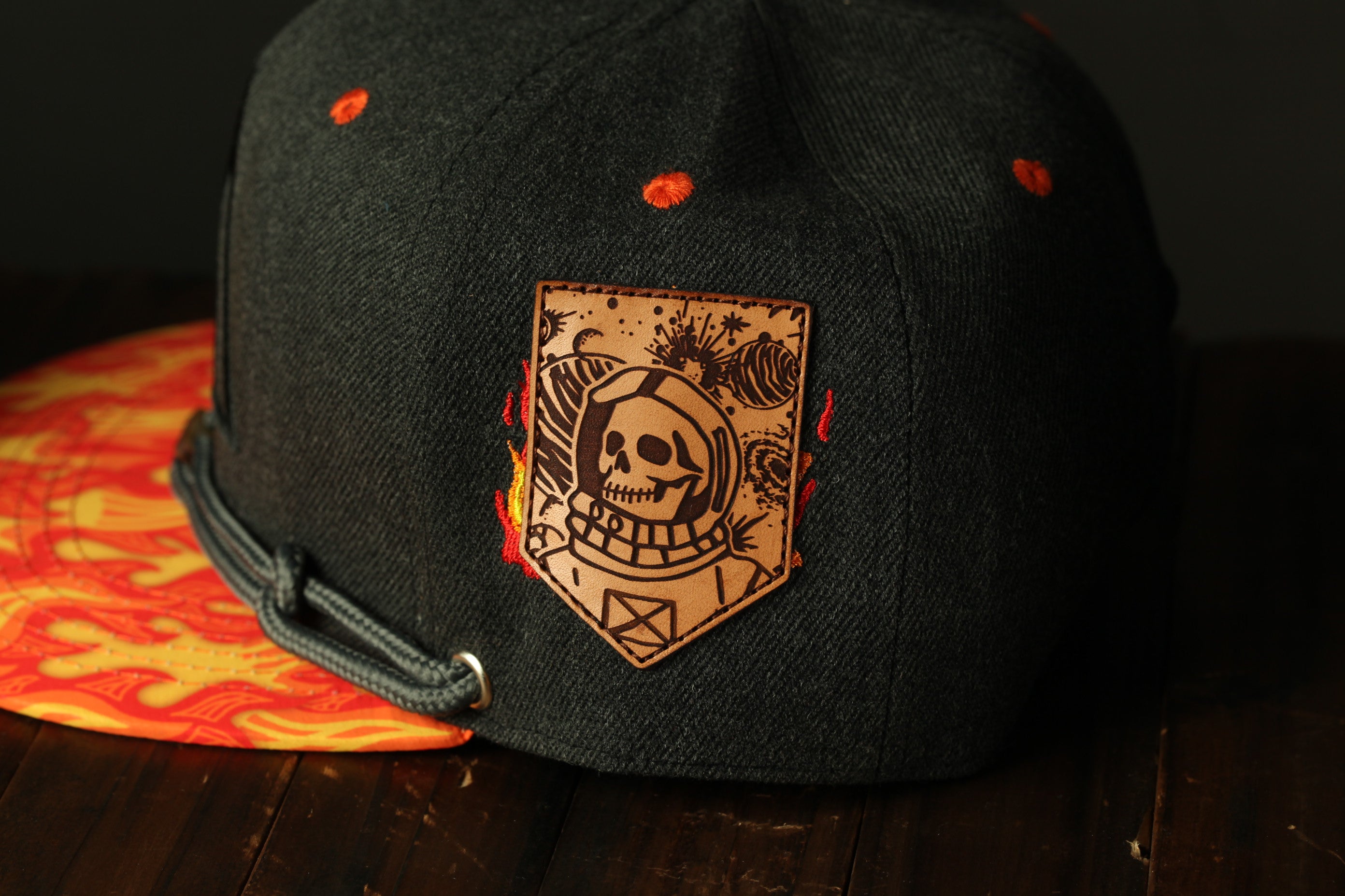 A black baseball cap with a leather patch featuring a skull and flames, placed on a wooden surface.