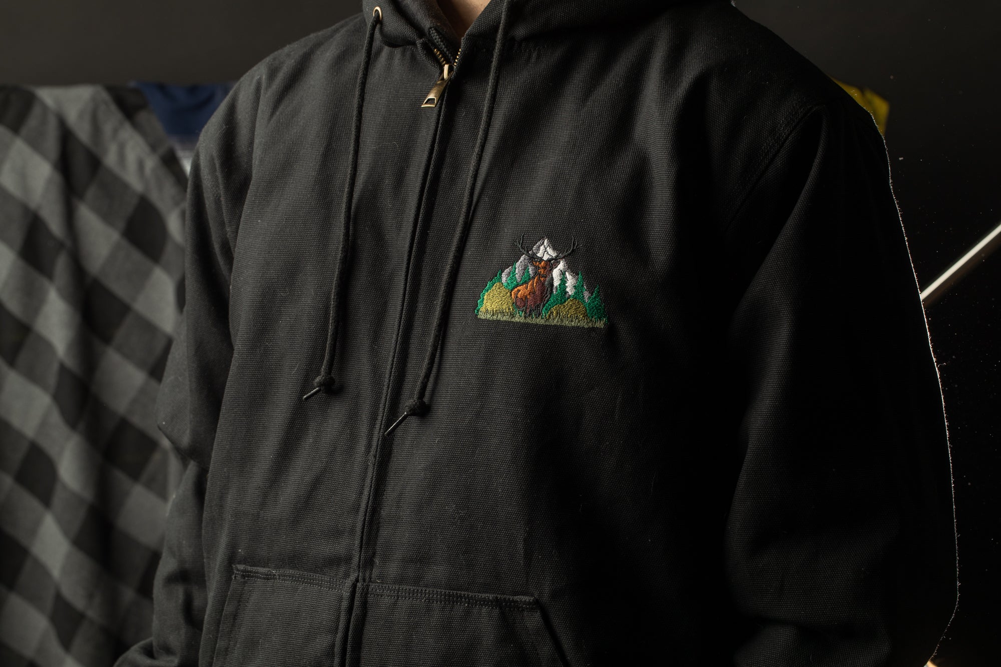 A close-up of a person wearing a black hooded sweatshirt with a mountain landscape embroidered on the front.