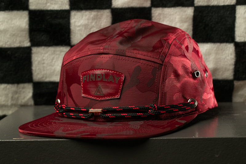 A red camouflage baseball cap with the word "FINDlay" printed on it, sitting on a black surface against a black and white checkered background.