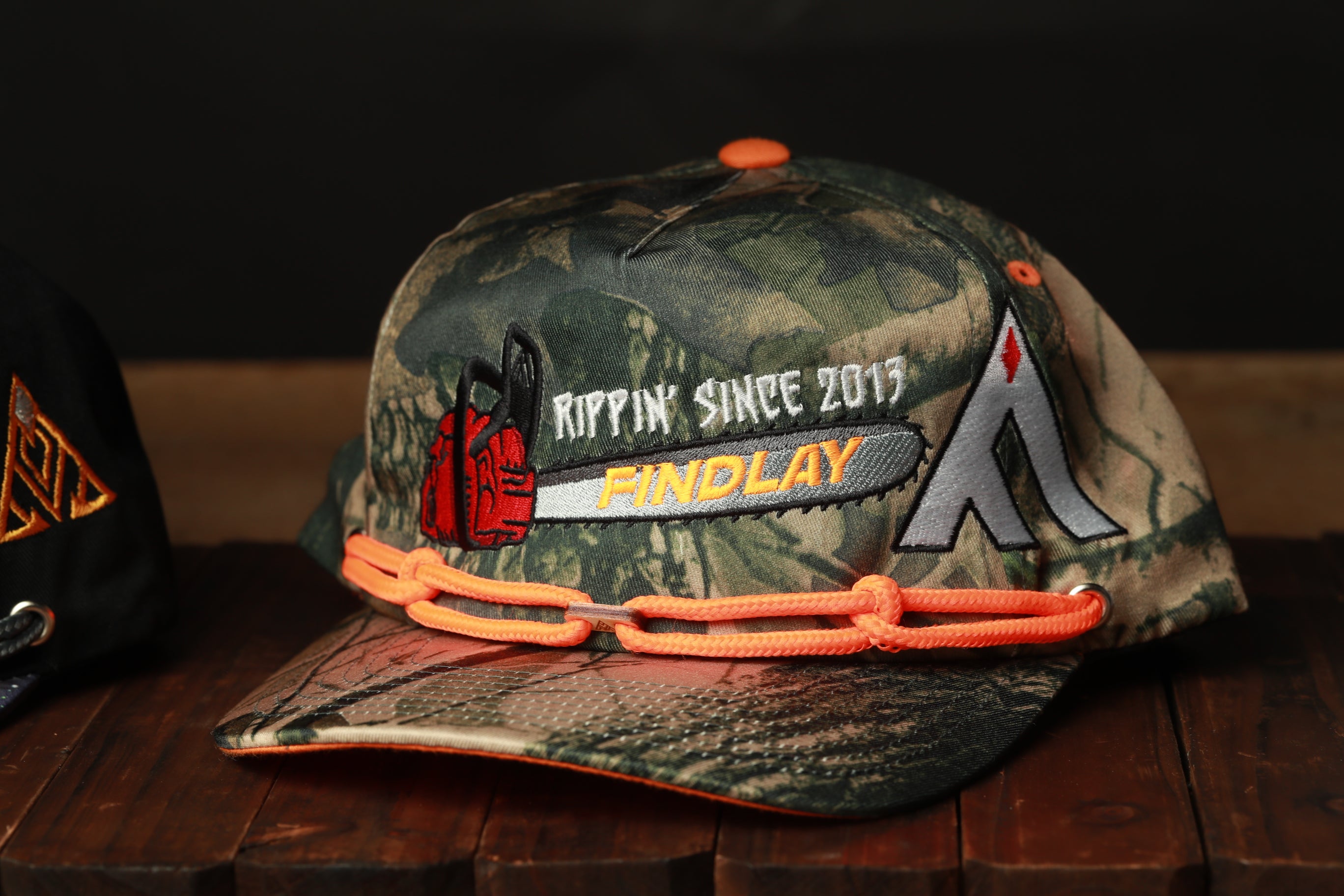A camouflage baseball cap with orange straps and a logo, sitting on a wooden surface.