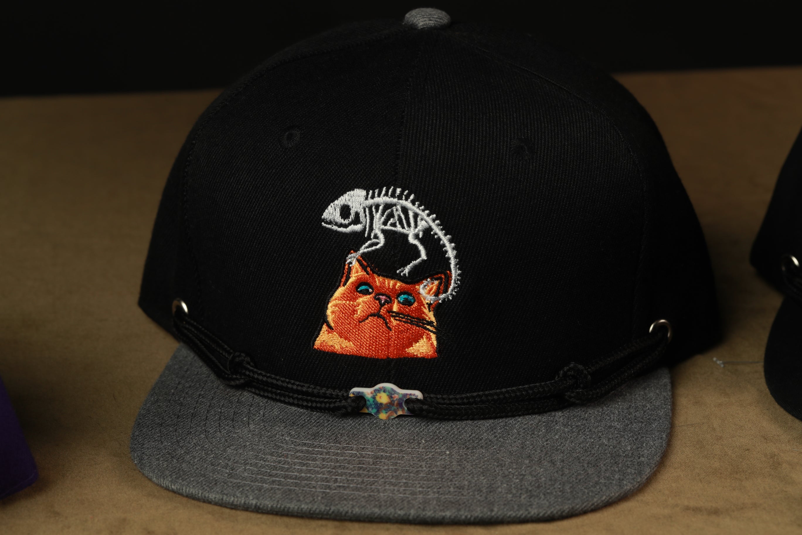 A black baseball cap with an embroidered image of a cat and a skeleton on the front.