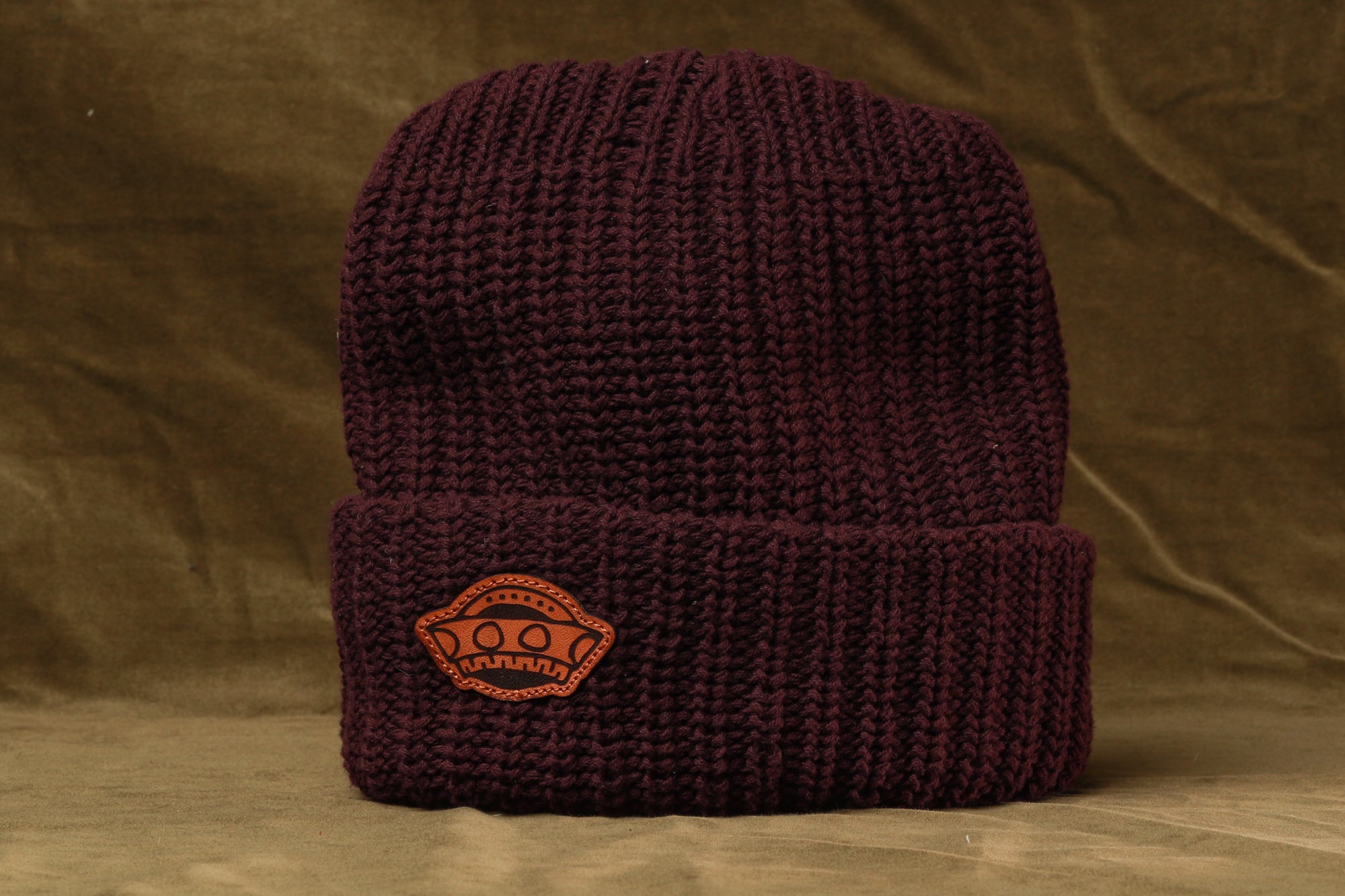 A burgundy knit beanie with a small orange patch on the front, sitting on a brown background.