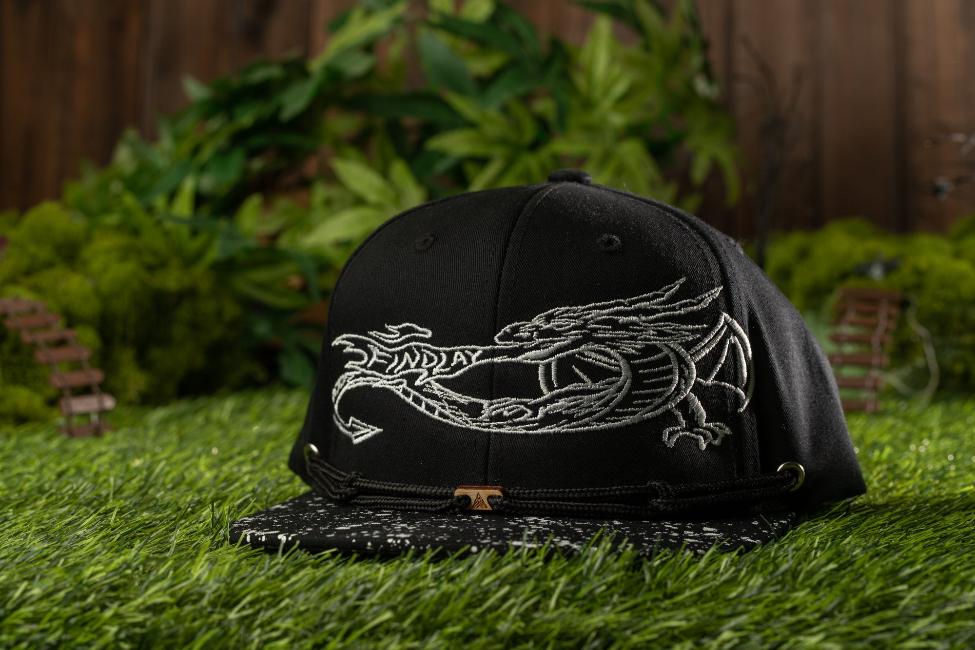 A black baseball cap with a white dragon design on it, placed on a grassy surface with some foliage in the background.