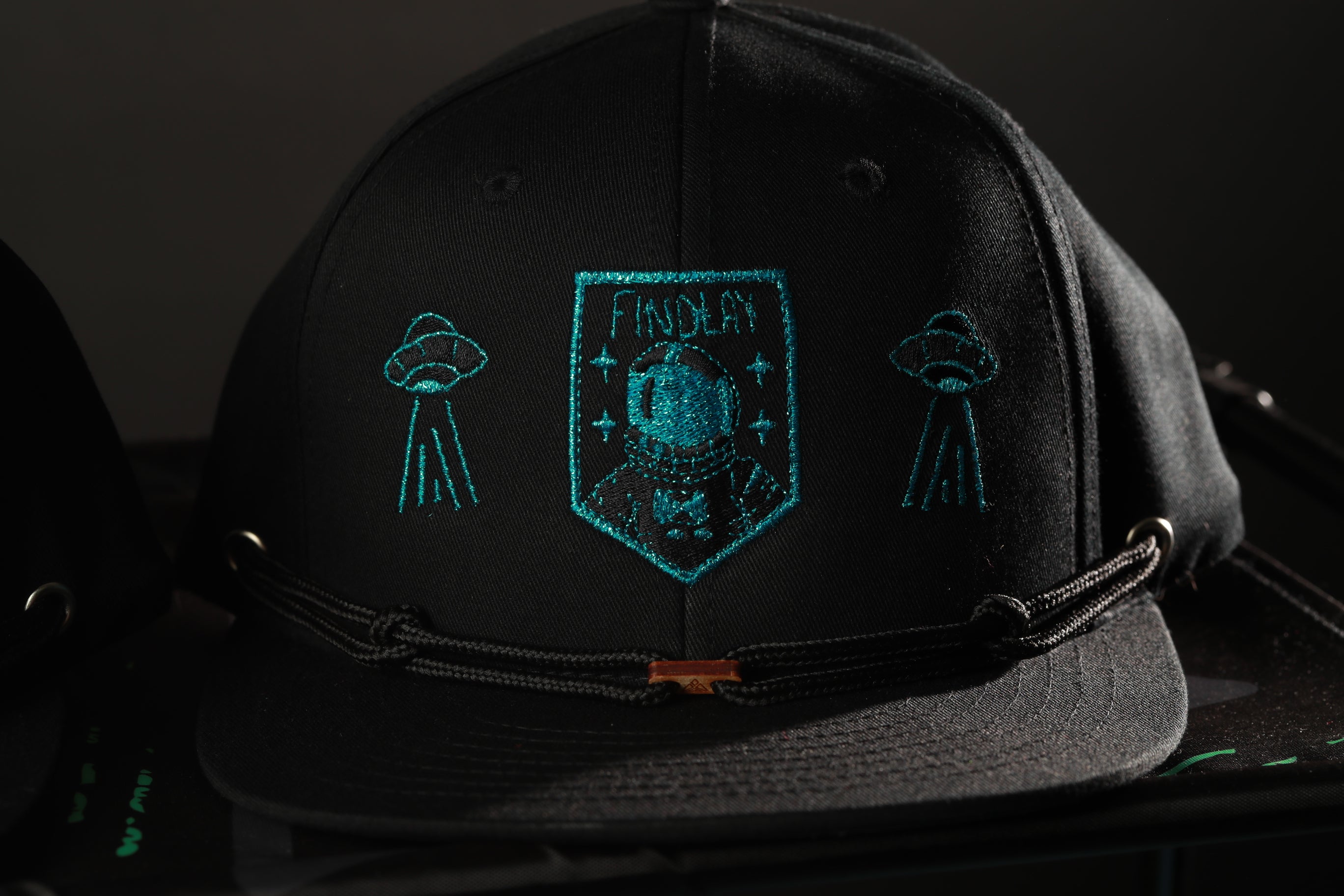 A black baseball cap with a teal embroidered design on the front, featuring a shield-shaped emblem with a person's face and the text "FINDAS".