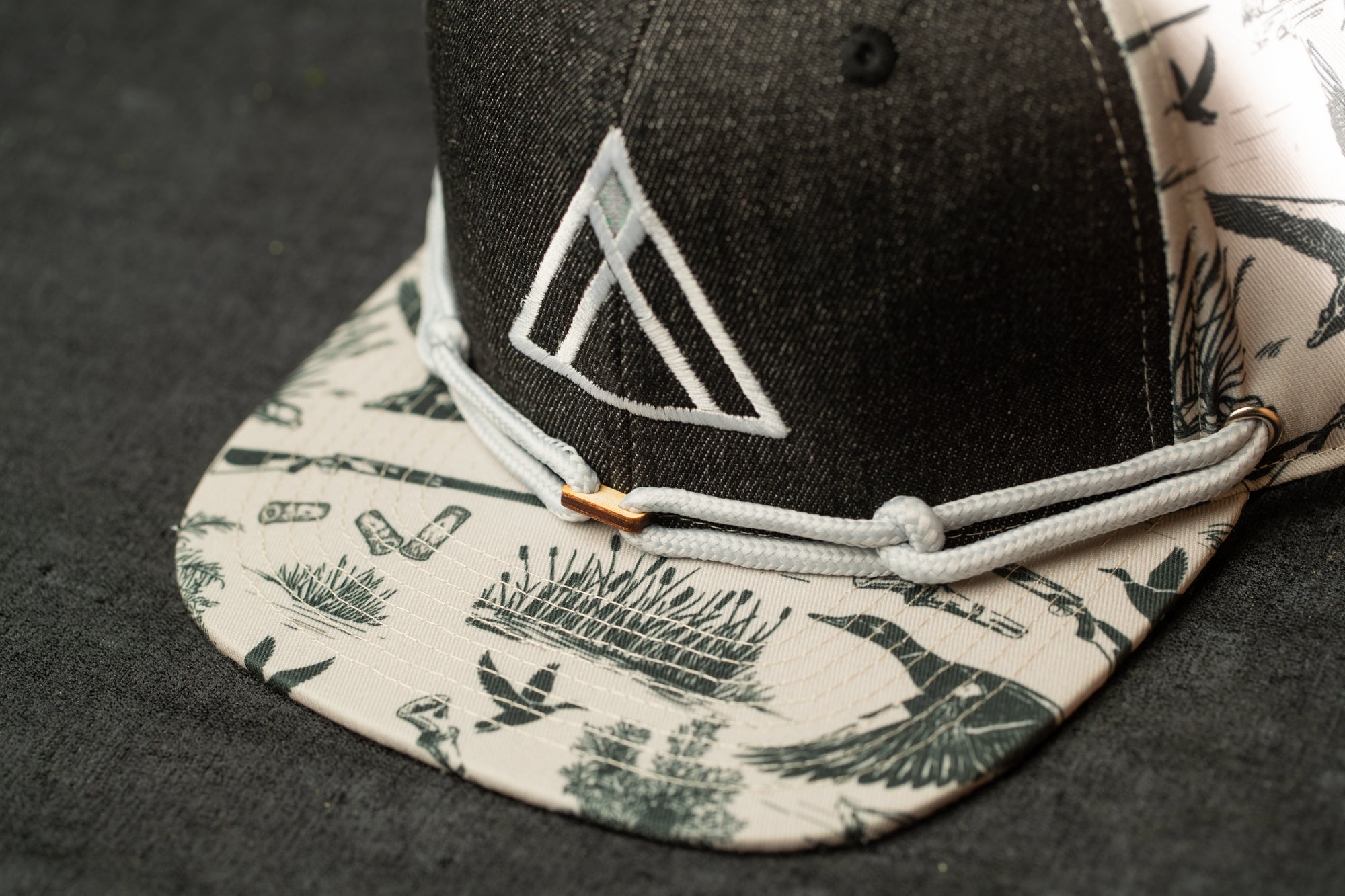 A close-up view of a black and white baseball cap with a pattern of ducks and other wildlife on the brim and a white triangular logo on the front.
