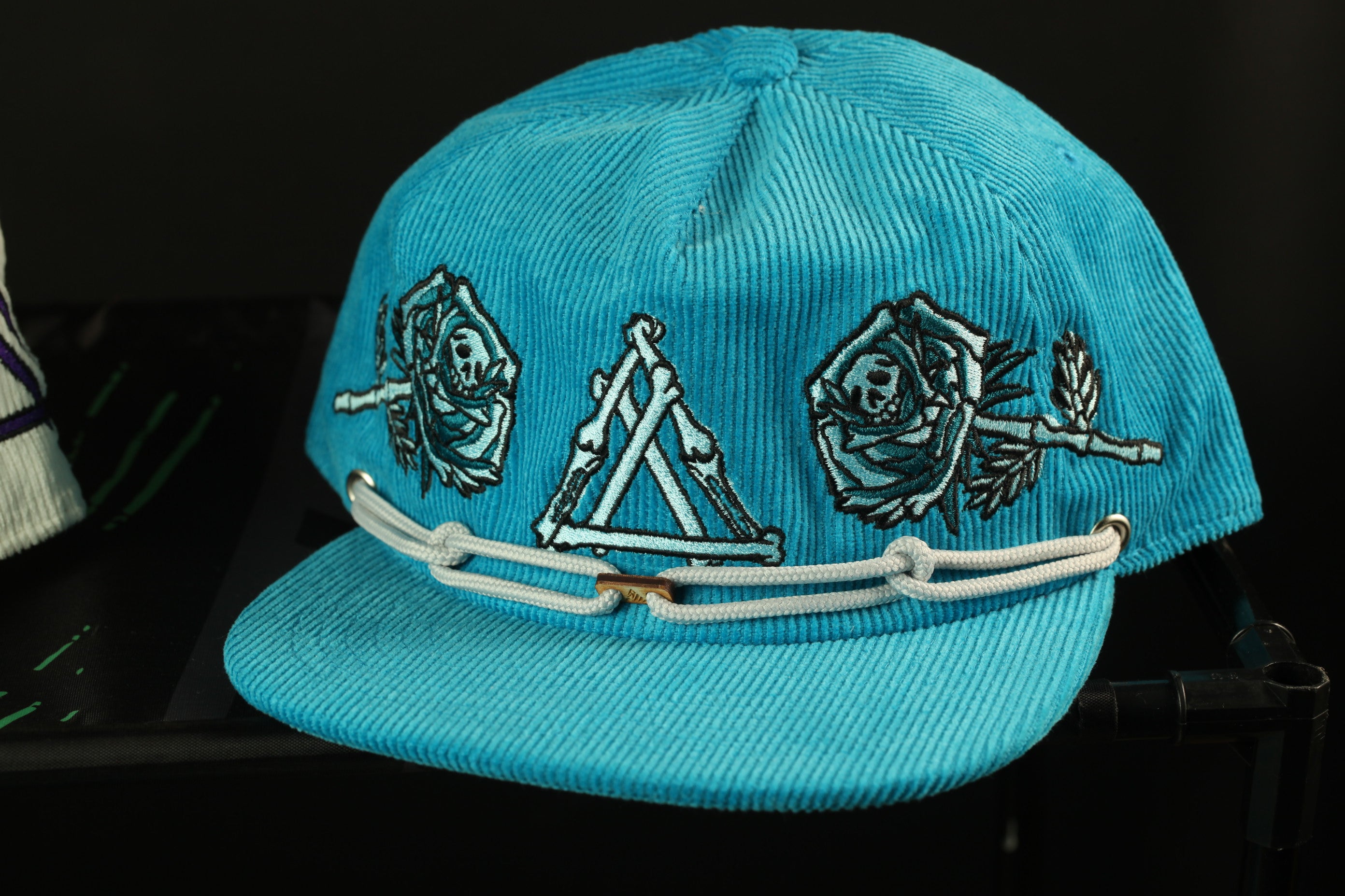 A turquoise baseball cap with a white lace trim and various decorative elements, including skull and crossbones symbols, is displayed against a dark background.