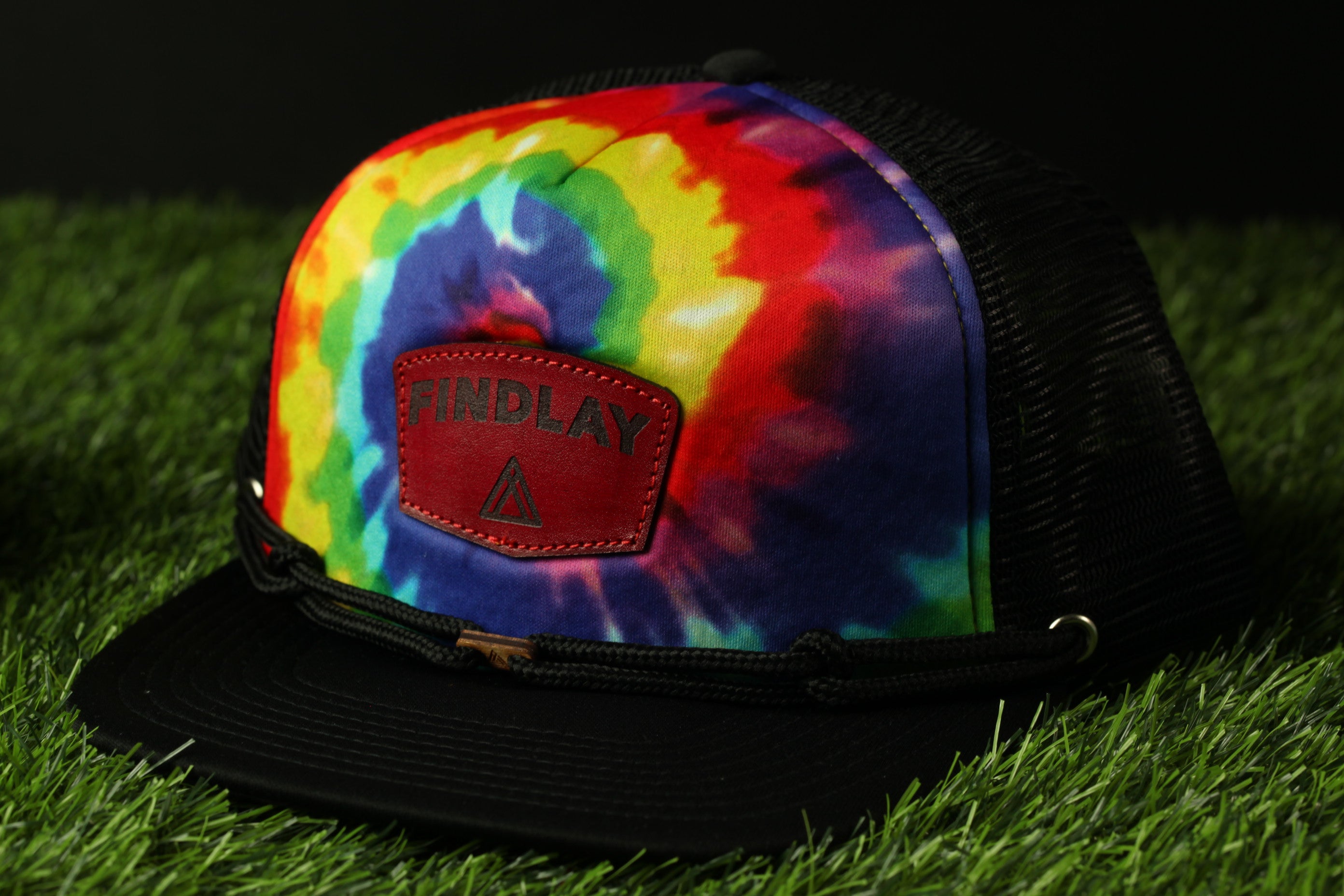 A colorful tie-dye baseball cap with the word "FINDlay" printed on it, placed on a grassy surface.