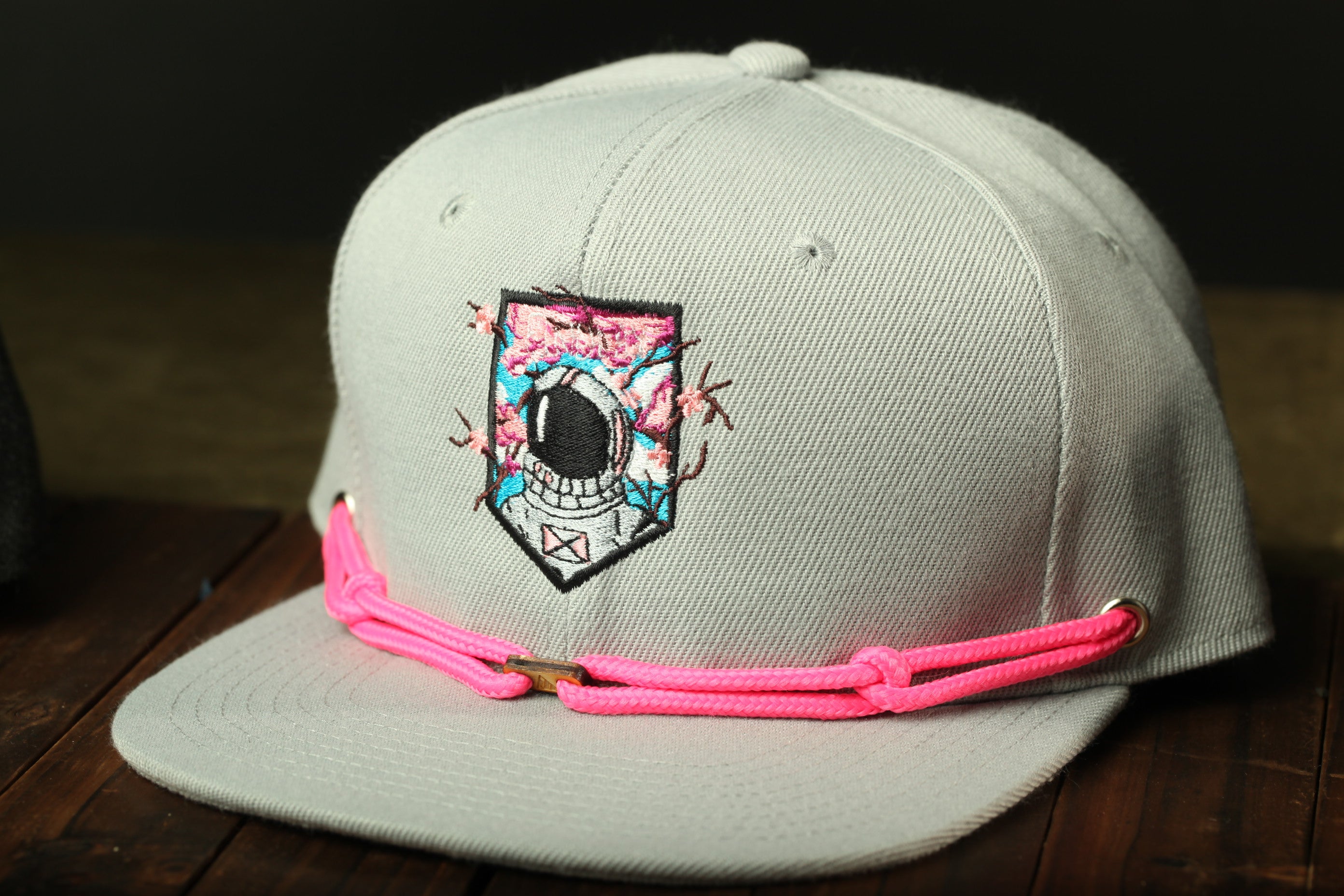 A white baseball cap with a colorful patch featuring a space helmet design, and a pink braided strap.