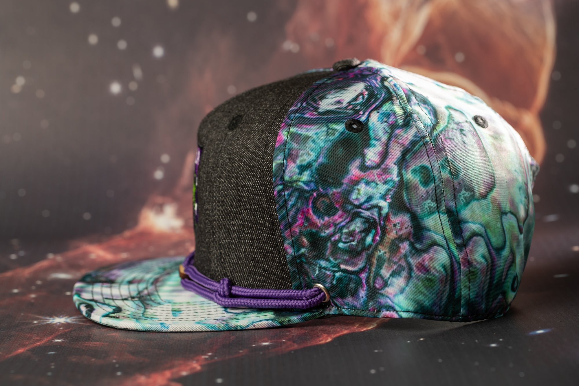 A colorful, patterned baseball cap with a black brim and purple strap is displayed against a dark background with a starry sky.