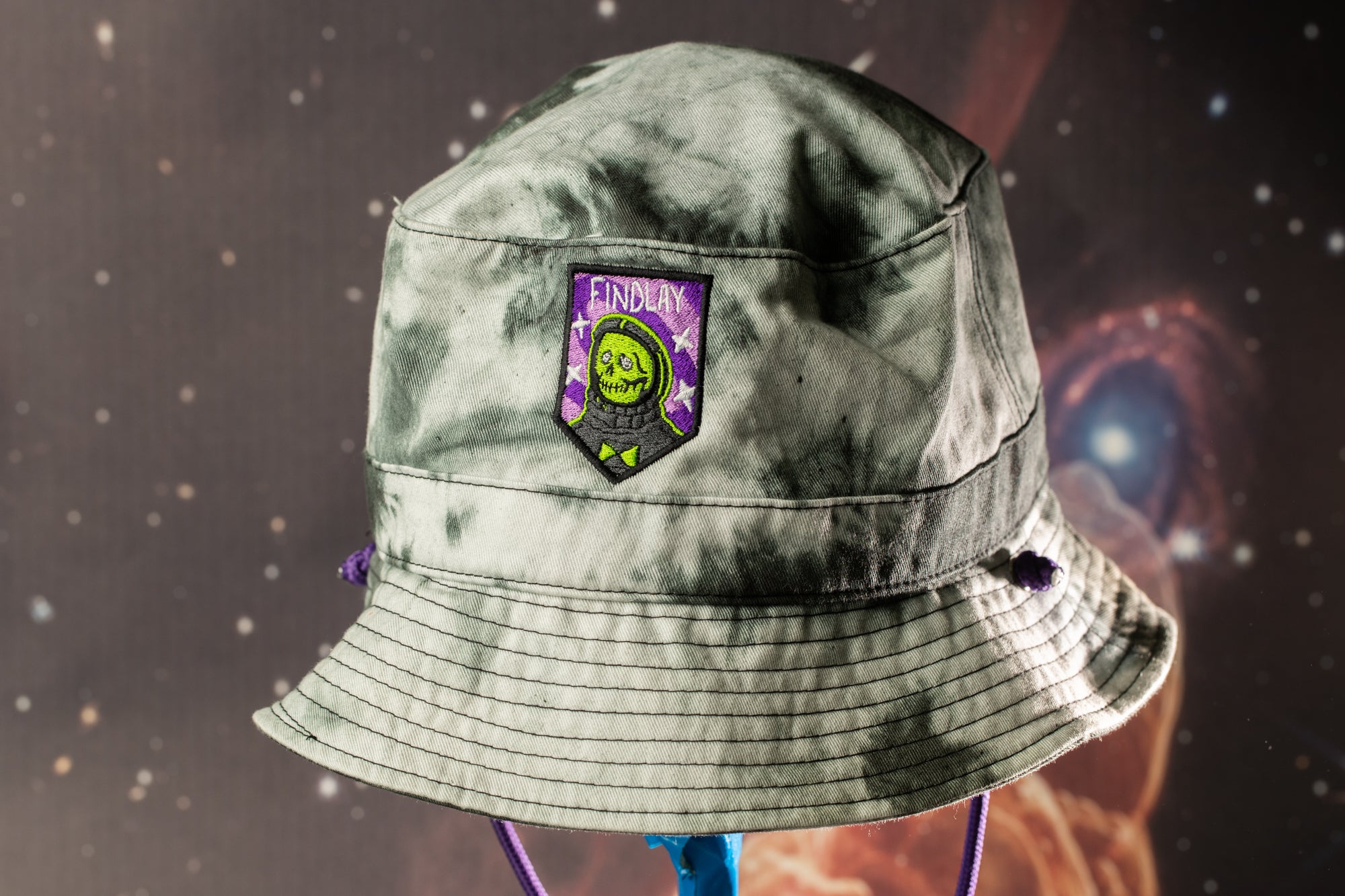 A gray and purple bucket hat with a green and purple patch on the front, featuring a skull and crossbones design.