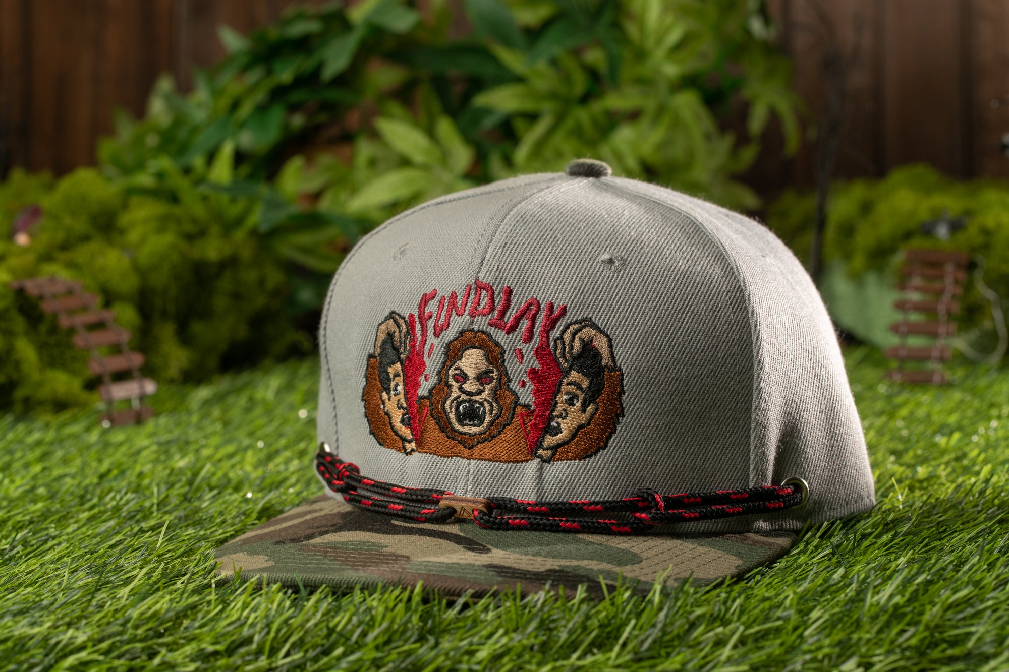 A gray baseball cap with a camouflage pattern and a graphic of a cartoon character is placed on a grassy surface.