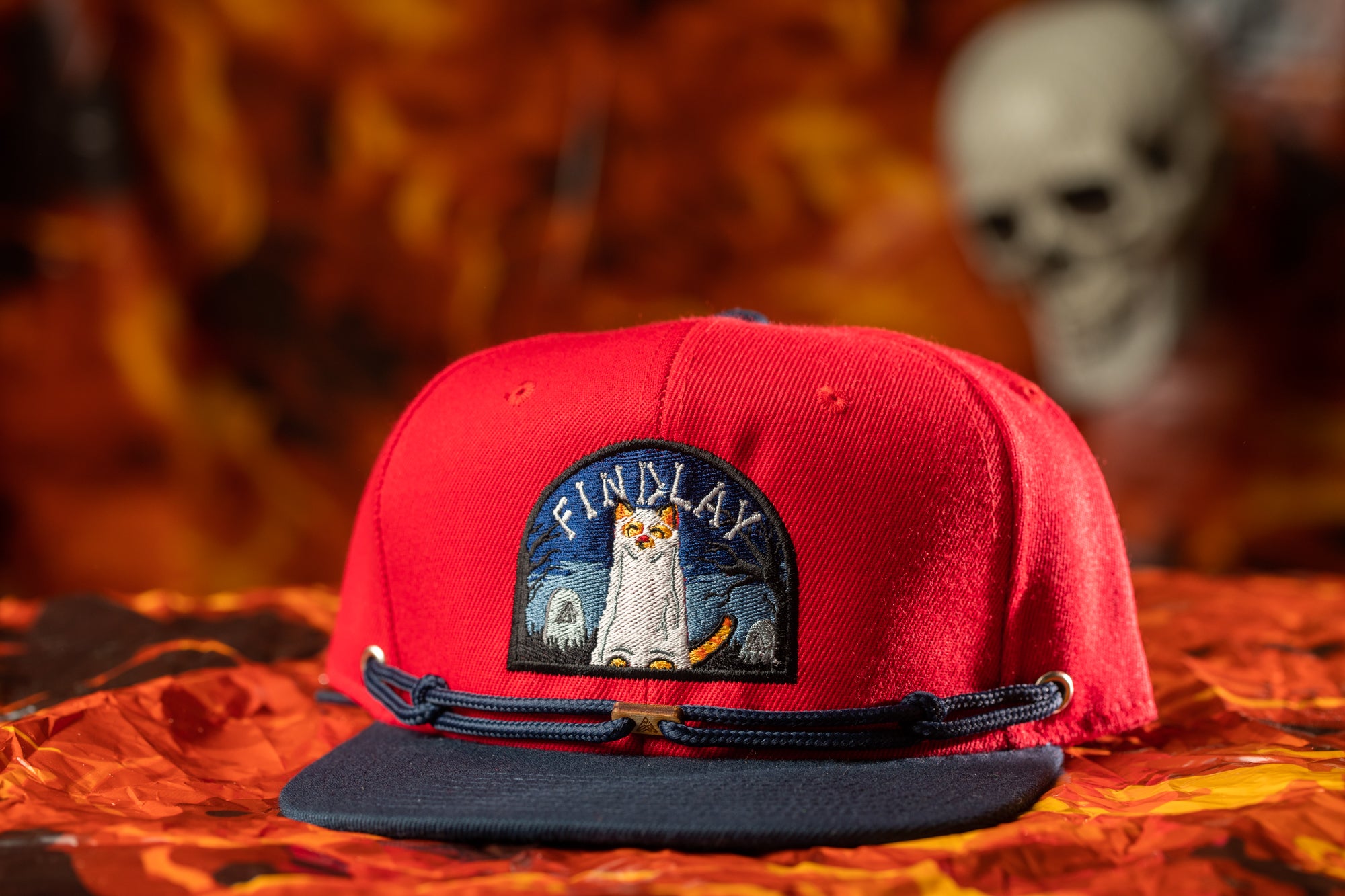 A red baseball cap with a cat embroidered on it, placed on a red and orange background.