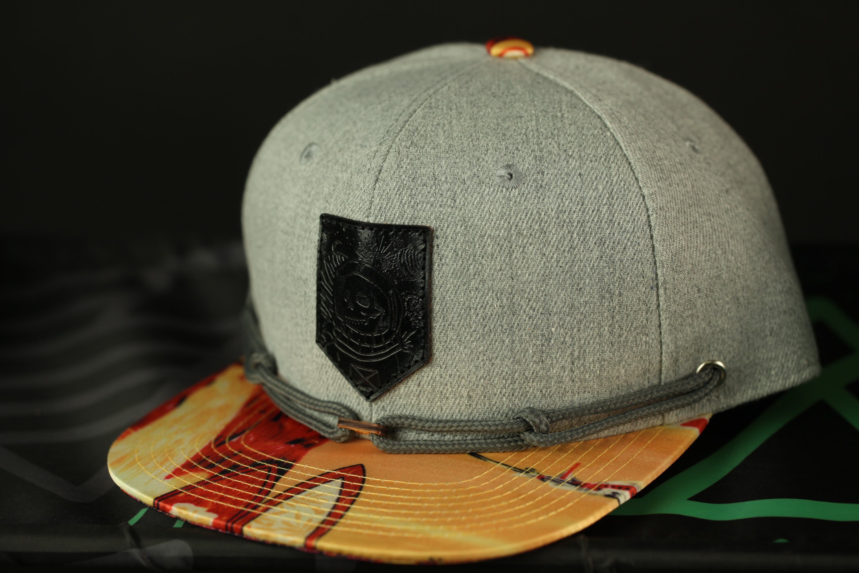 A gray baseball cap with a black patch and a colorful printed brim is displayed against a black background.