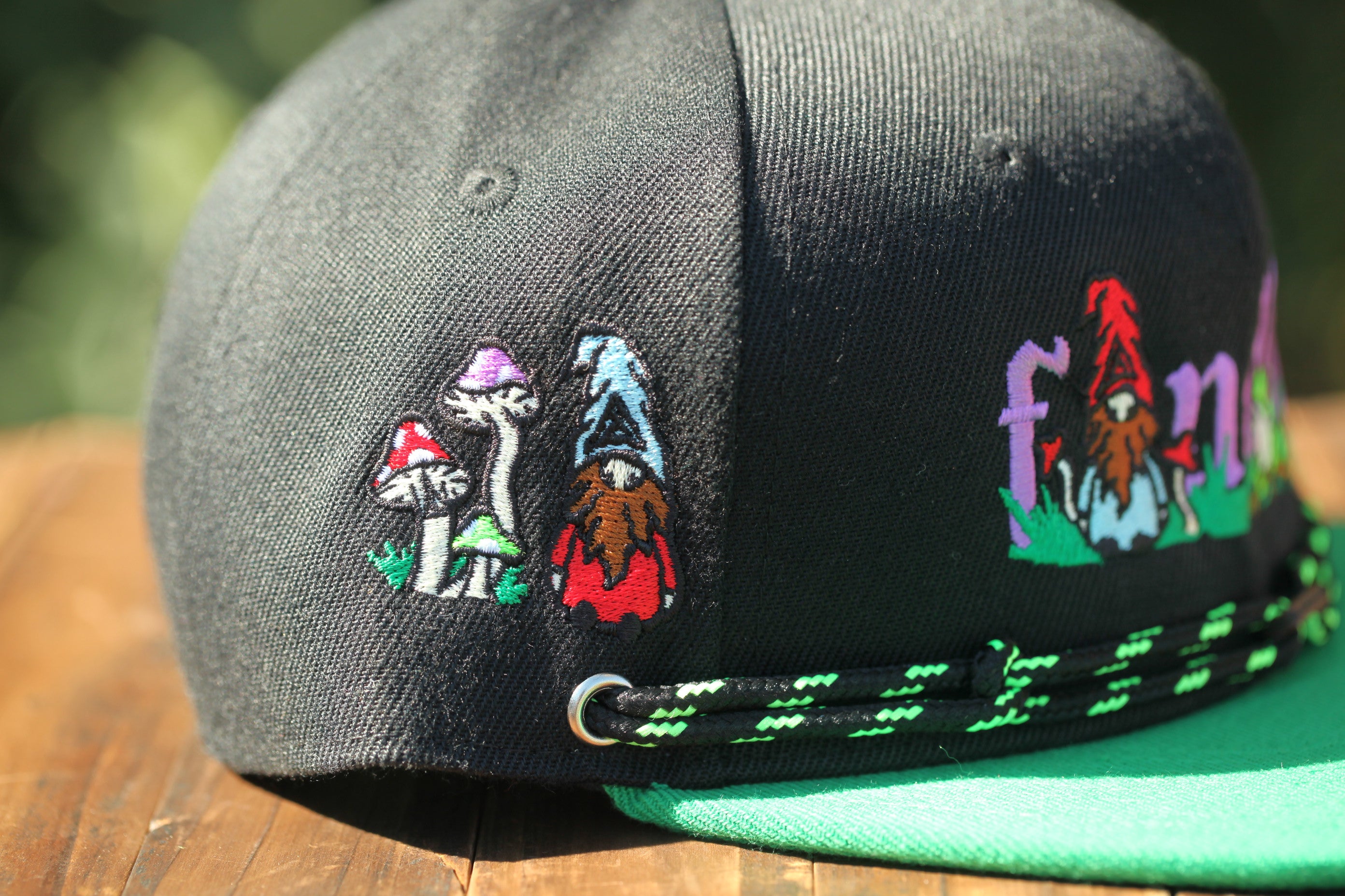 A close-up view of a black baseball cap with colorful embroidered designs, including mushrooms and gnomes, on the front.
