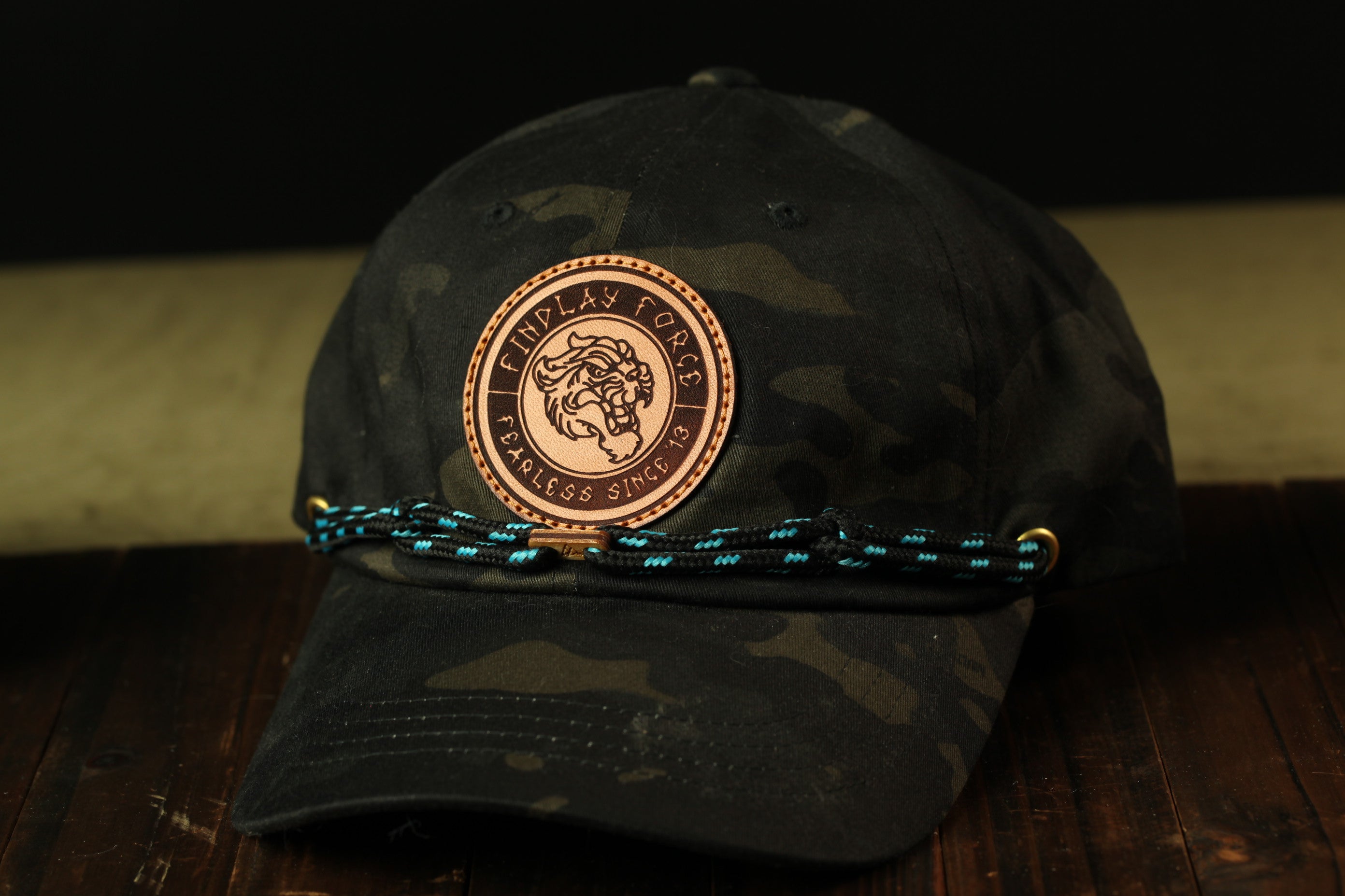 A black camouflage baseball cap with a circular patch on the front, featuring a lion's head design, and a blue braided strap.