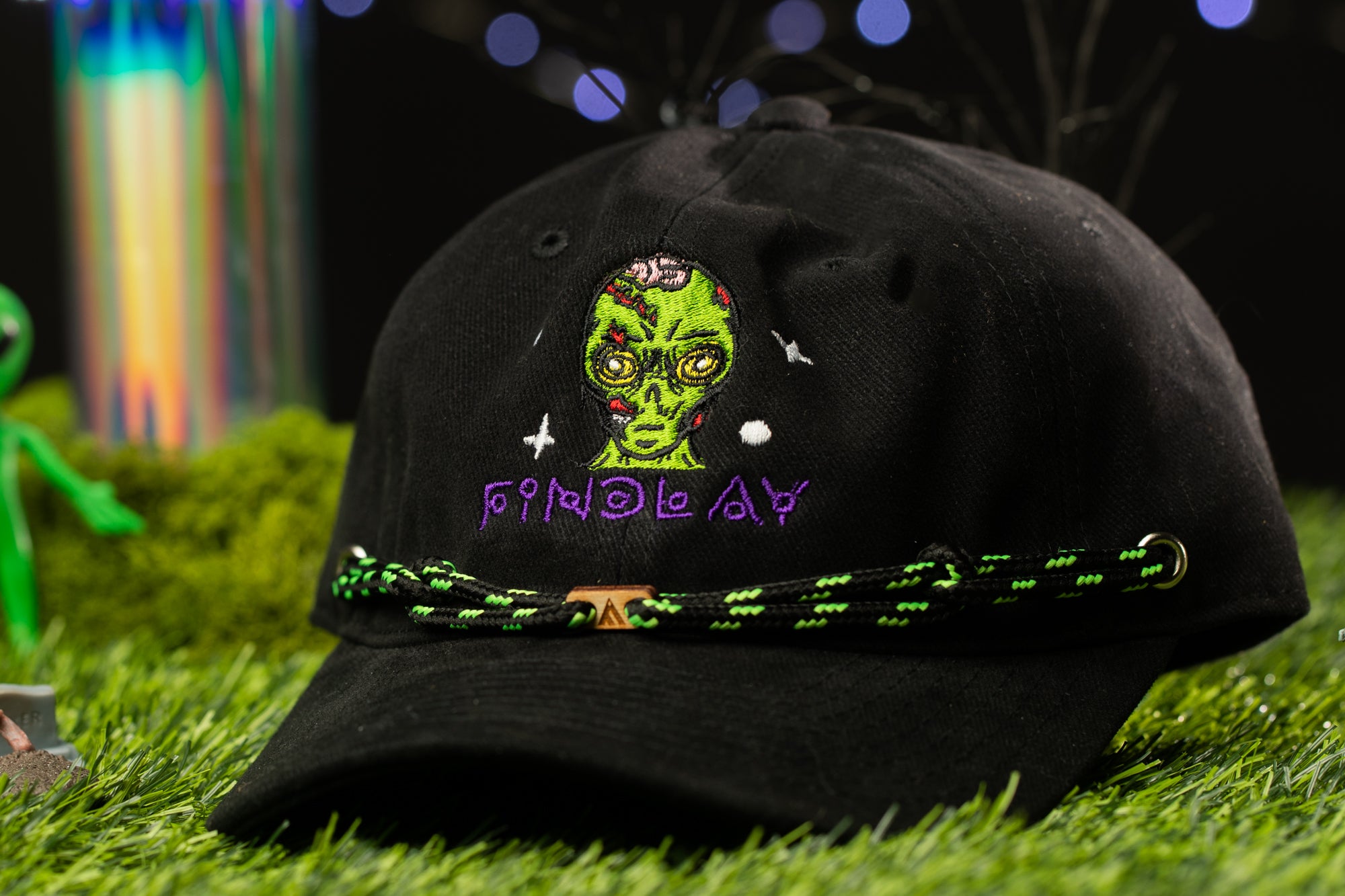A black baseball cap with a green alien face design and purple text on the front, placed on a grassy surface with colorful lights in the background.