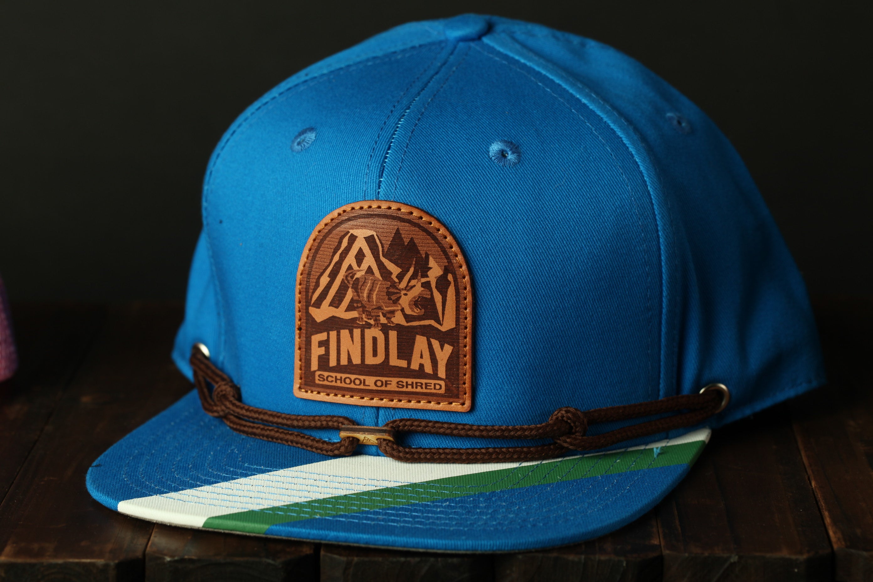 A blue baseball cap with a brown leather patch that says "FINDLAY" and "SCHOOL OF SHRED".