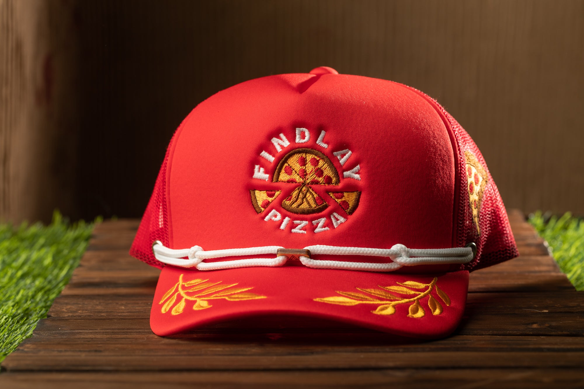 A red baseball cap with the logo of Findlay Pizza on it, placed on a wooden surface with grass visible in the background.
