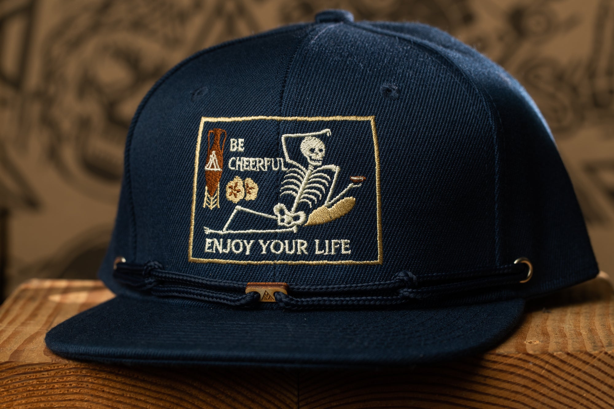 A navy blue baseball cap with a white and gold embroidered patch on the front, featuring a skeleton and the text "Be cheerful, enjoy your life".