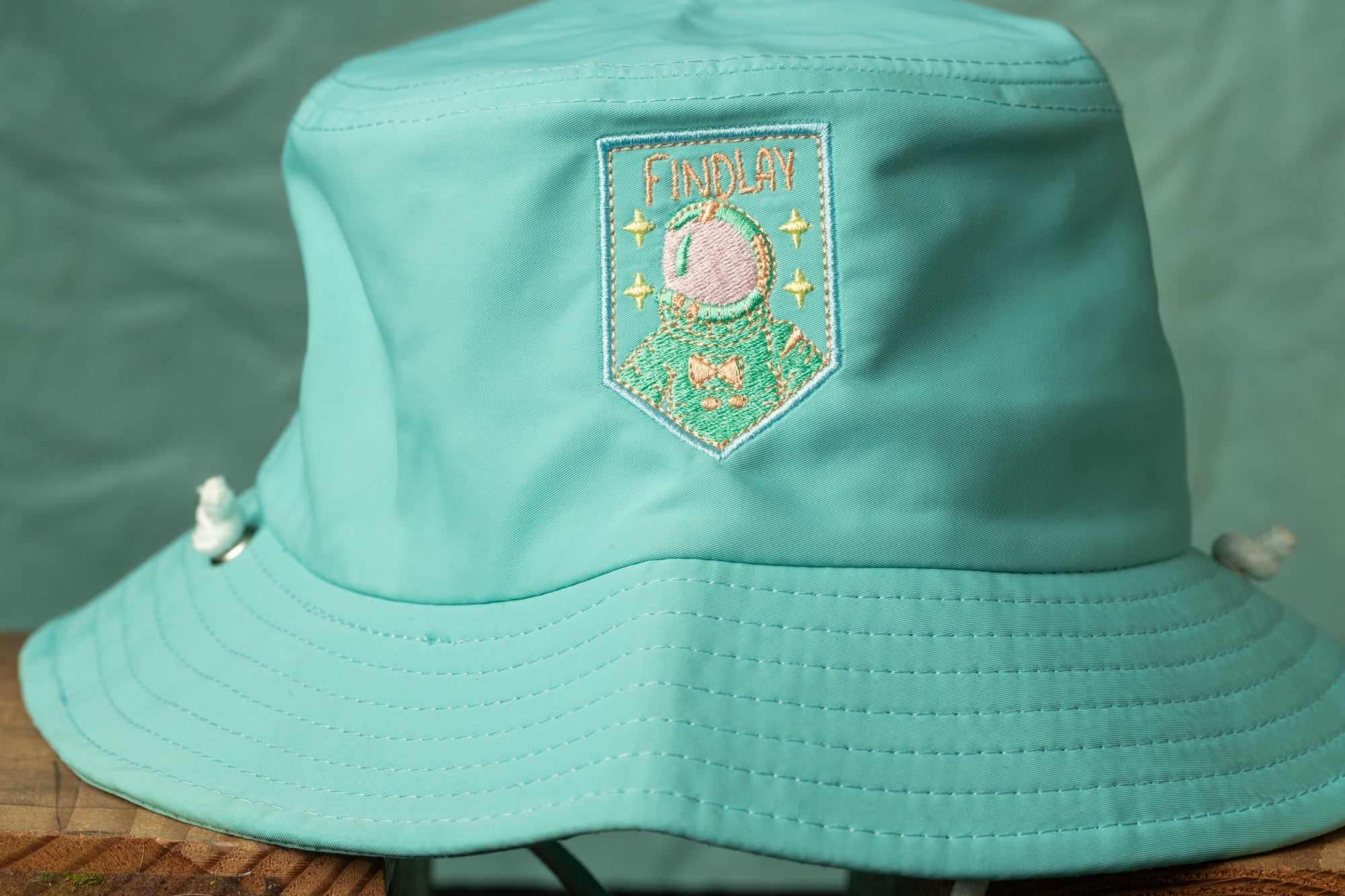 A turquoise bucket hat with a patch featuring an astronaut and the word "FINDLAY" printed on it.