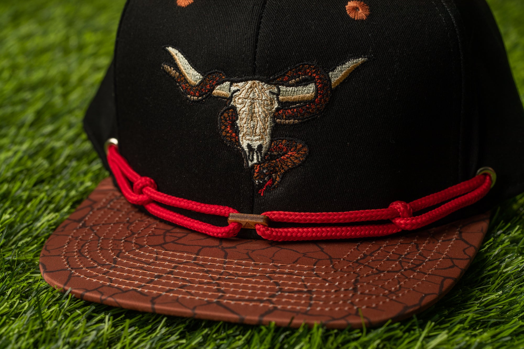 A close-up view of a black baseball cap with a red braided strap and an embroidered design of a bull's head and snake on the front.