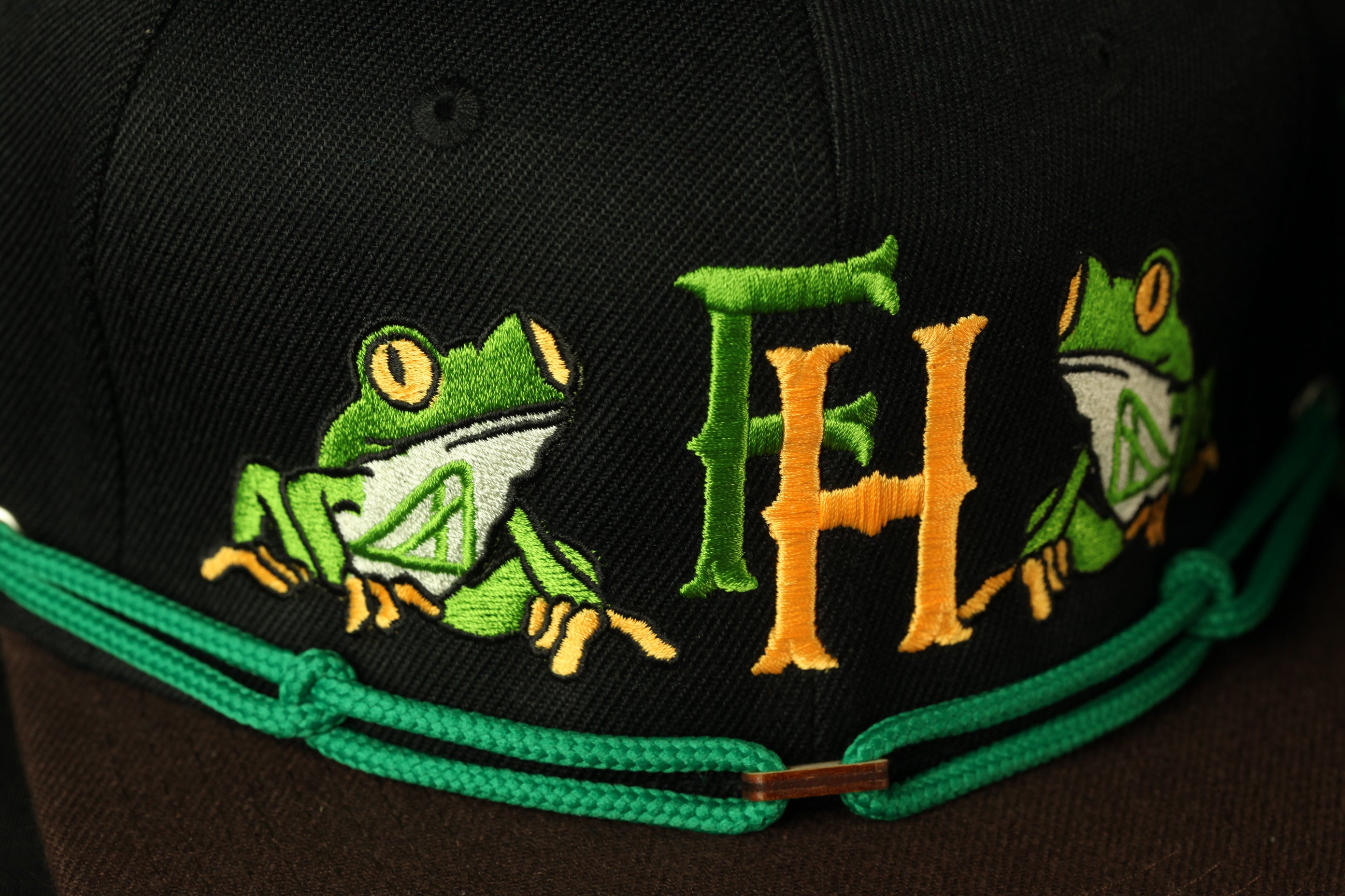A close-up of a black baseball cap with a green and orange embroidered design featuring two green frogs.