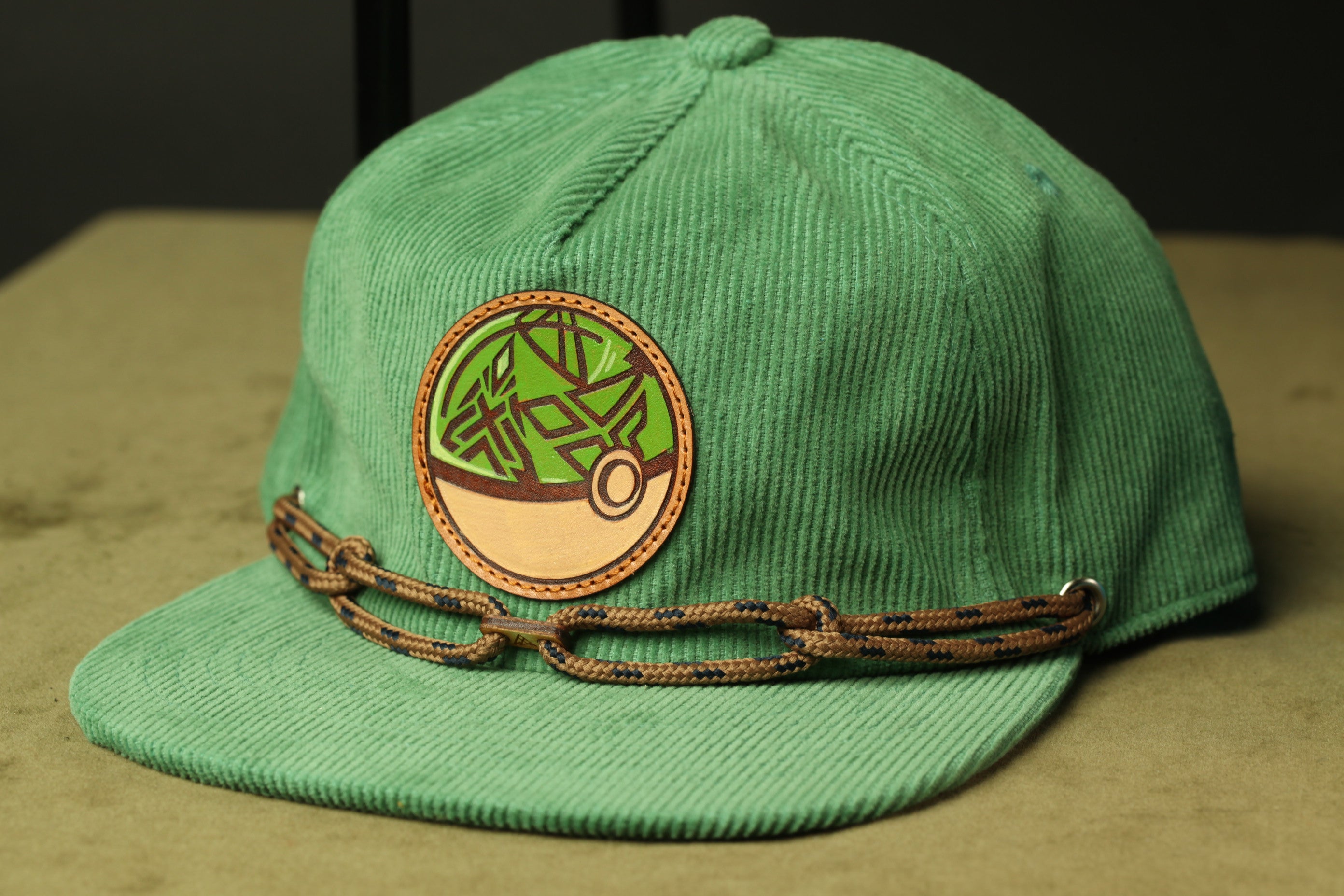 A green baseball cap with a circular patch featuring a tree design and a brown chain strap.