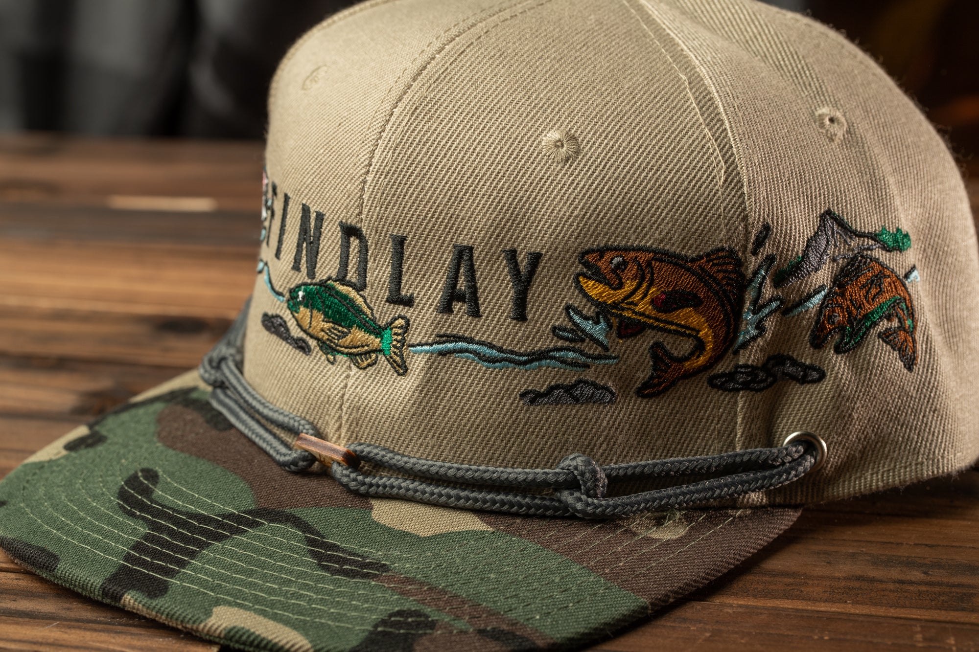 A beige baseball cap with a camouflage pattern and embroidered fish designs on the front.