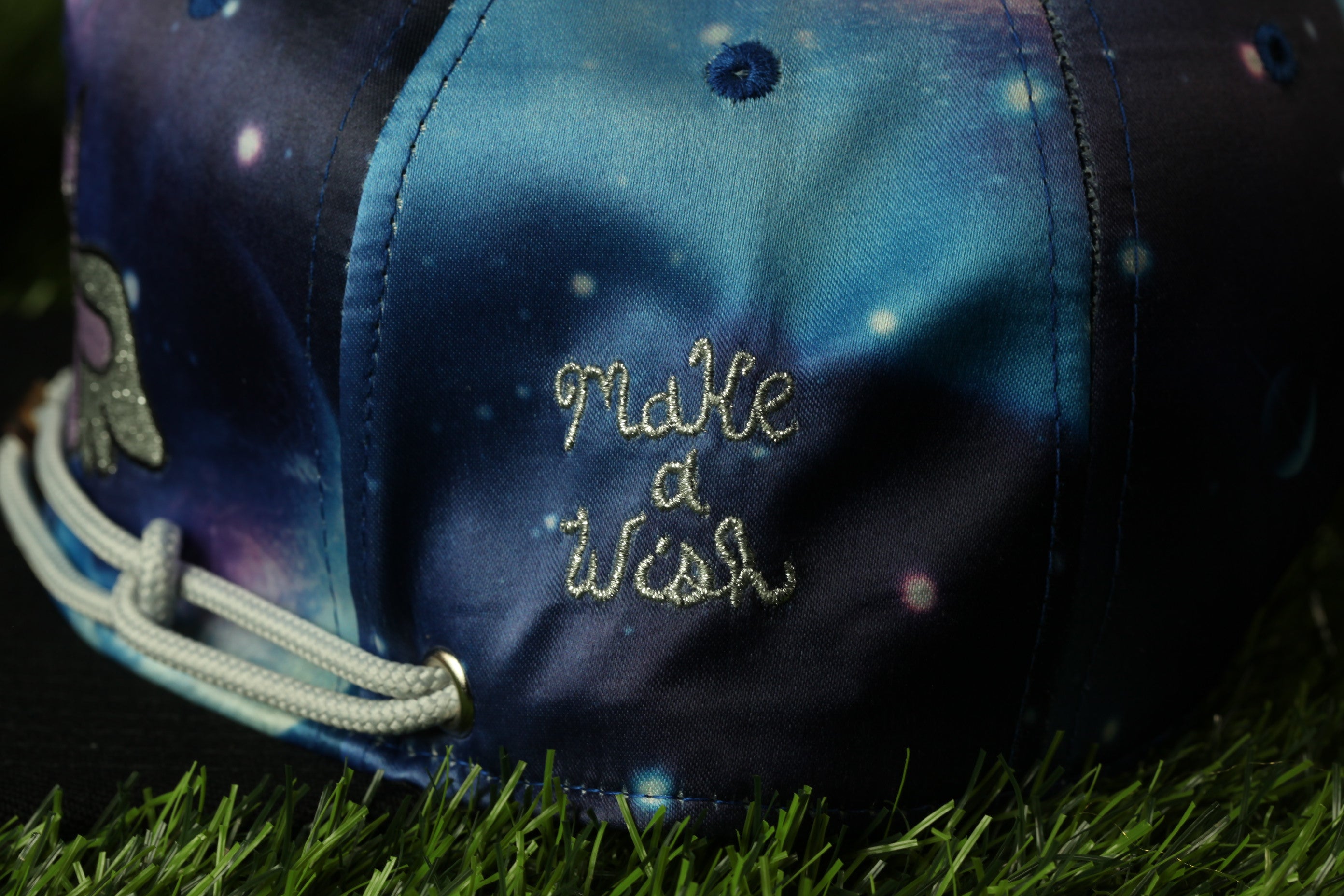 A close-up of a colorful baseball cap with the embroidered phrase "Make a wish" on it, resting on a grassy surface.