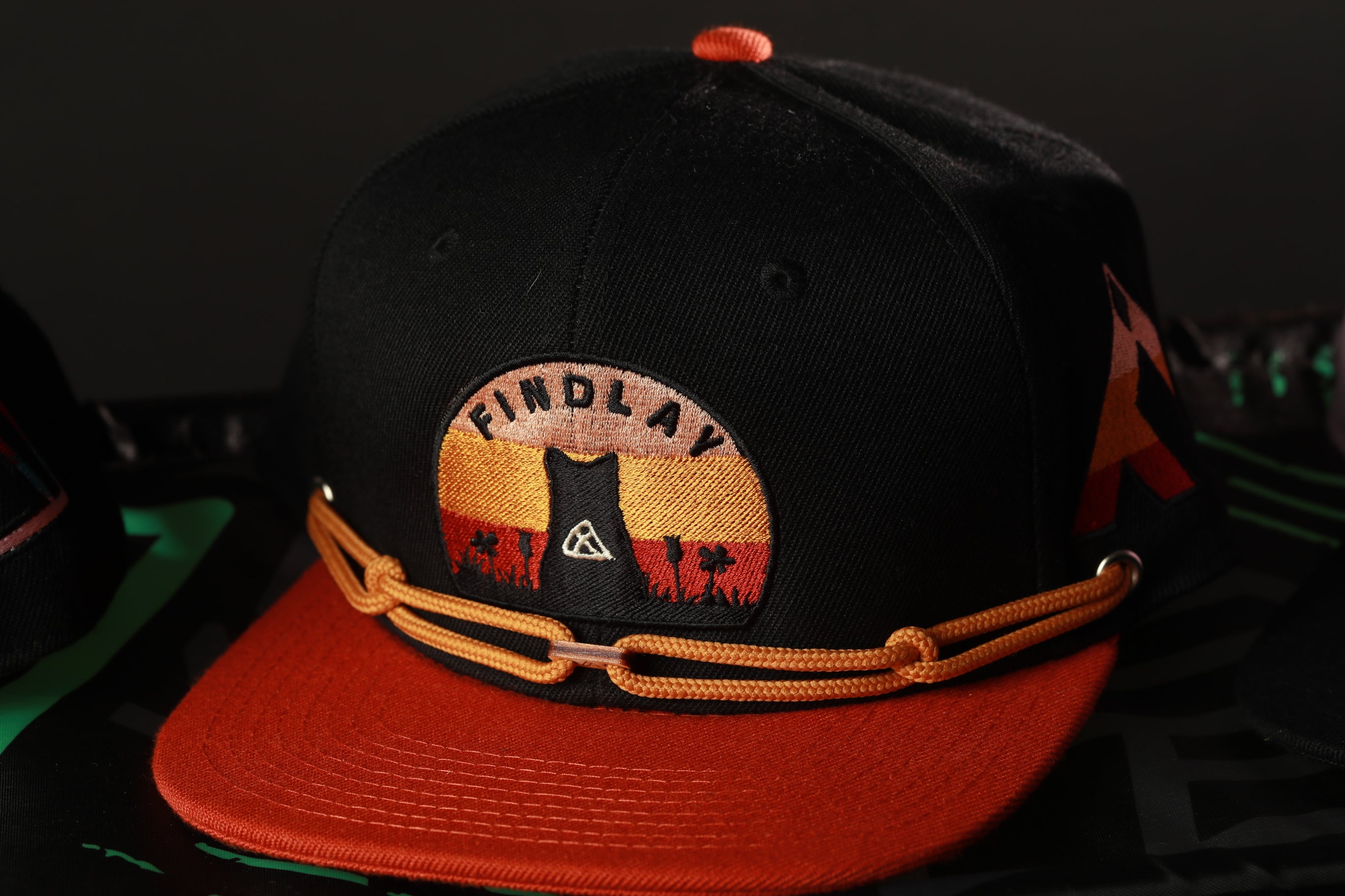 A black baseball cap with an orange brim and a patch on the front, featuring the word "FINDLAY" and a graphic design.