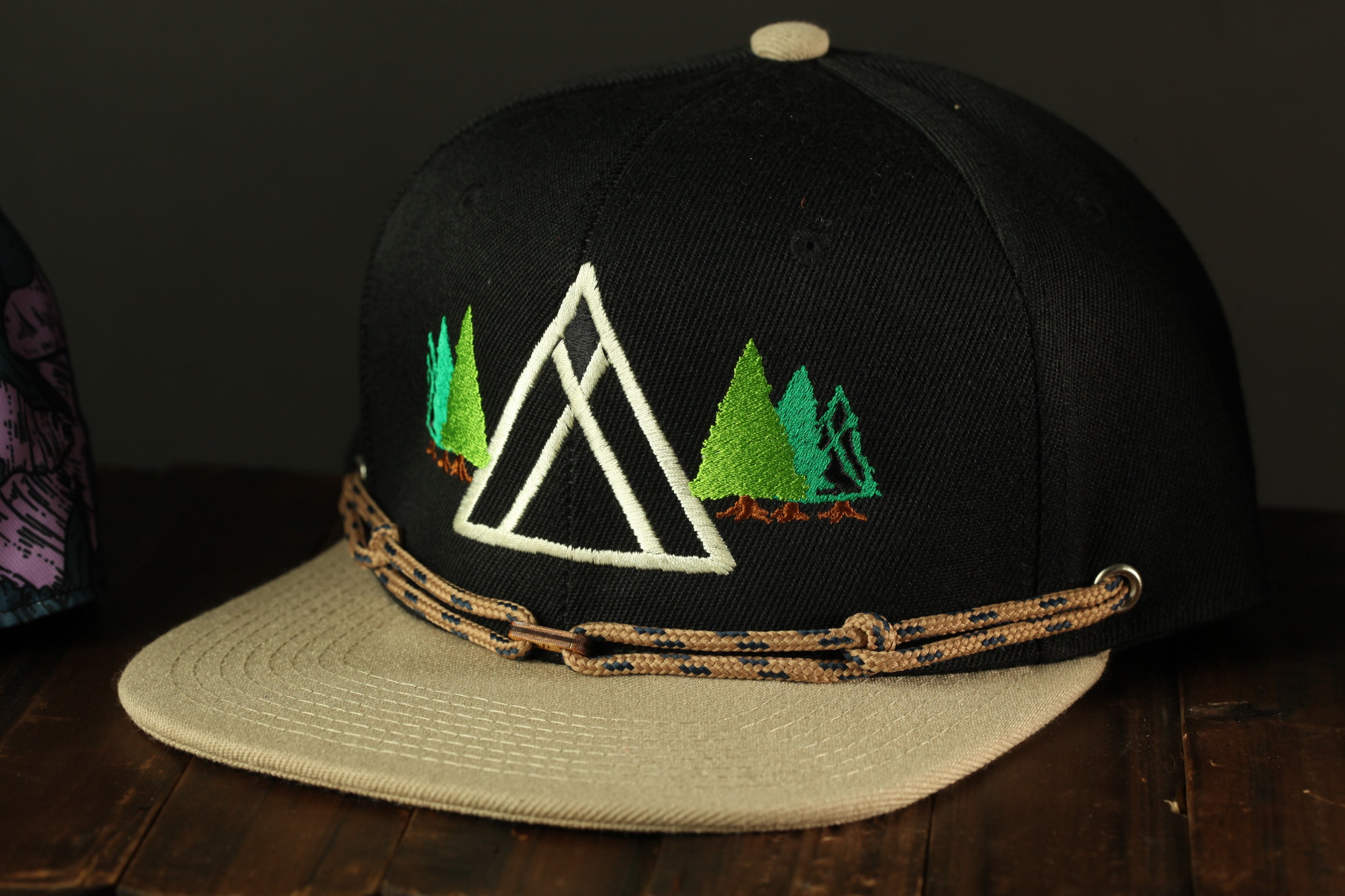 A black baseball cap with a beige brim and a white triangle design on the front, featuring a forest scene.