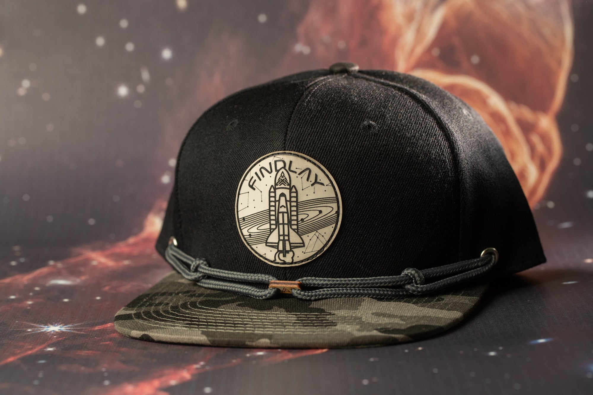 A black baseball cap with a space shuttle design on it, placed against a background of a space-themed image.