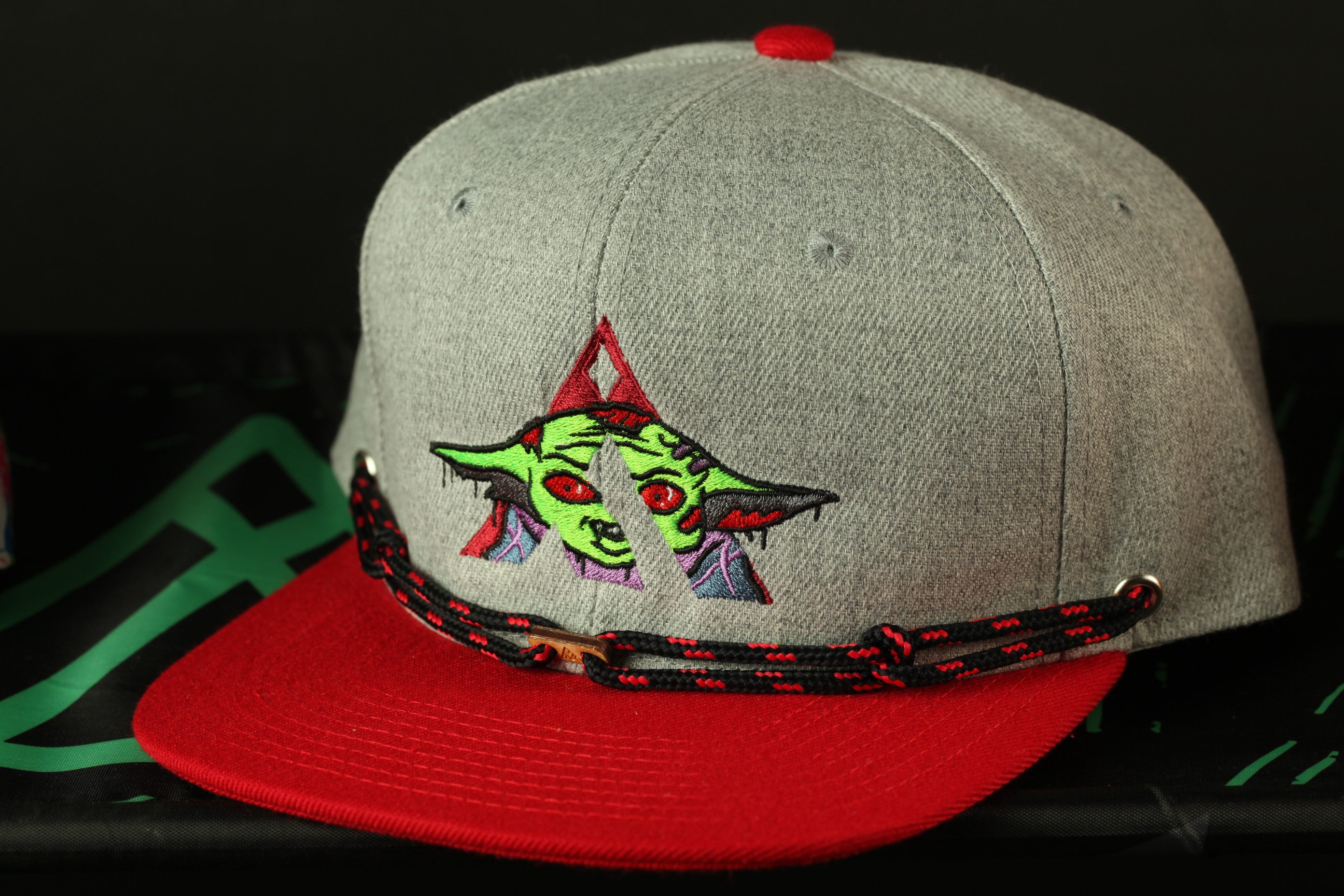 A gray baseball cap with a red brim and a graphic of a green alien-like creature on the front.
