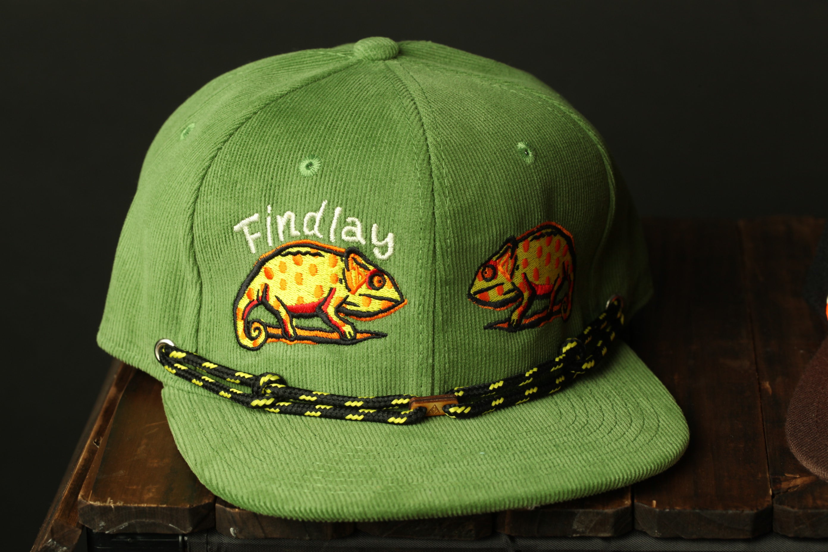 A green baseball cap with a black and yellow braided strap, featuring two chameleon illustrations on the front.
