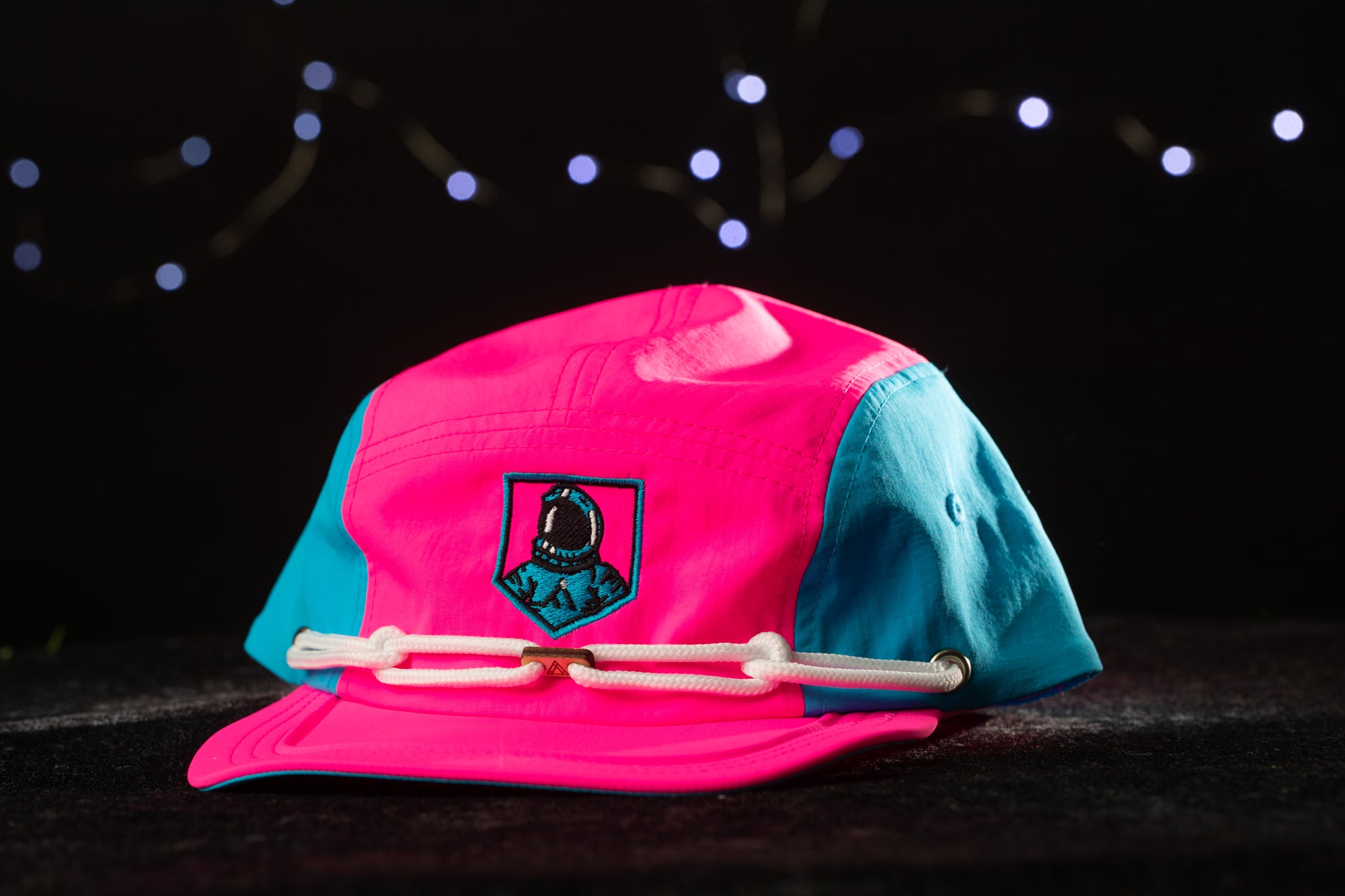 A pink and blue baseball cap with a small embroidered patch on the front, placed on a dark surface against a background of twinkling lights.