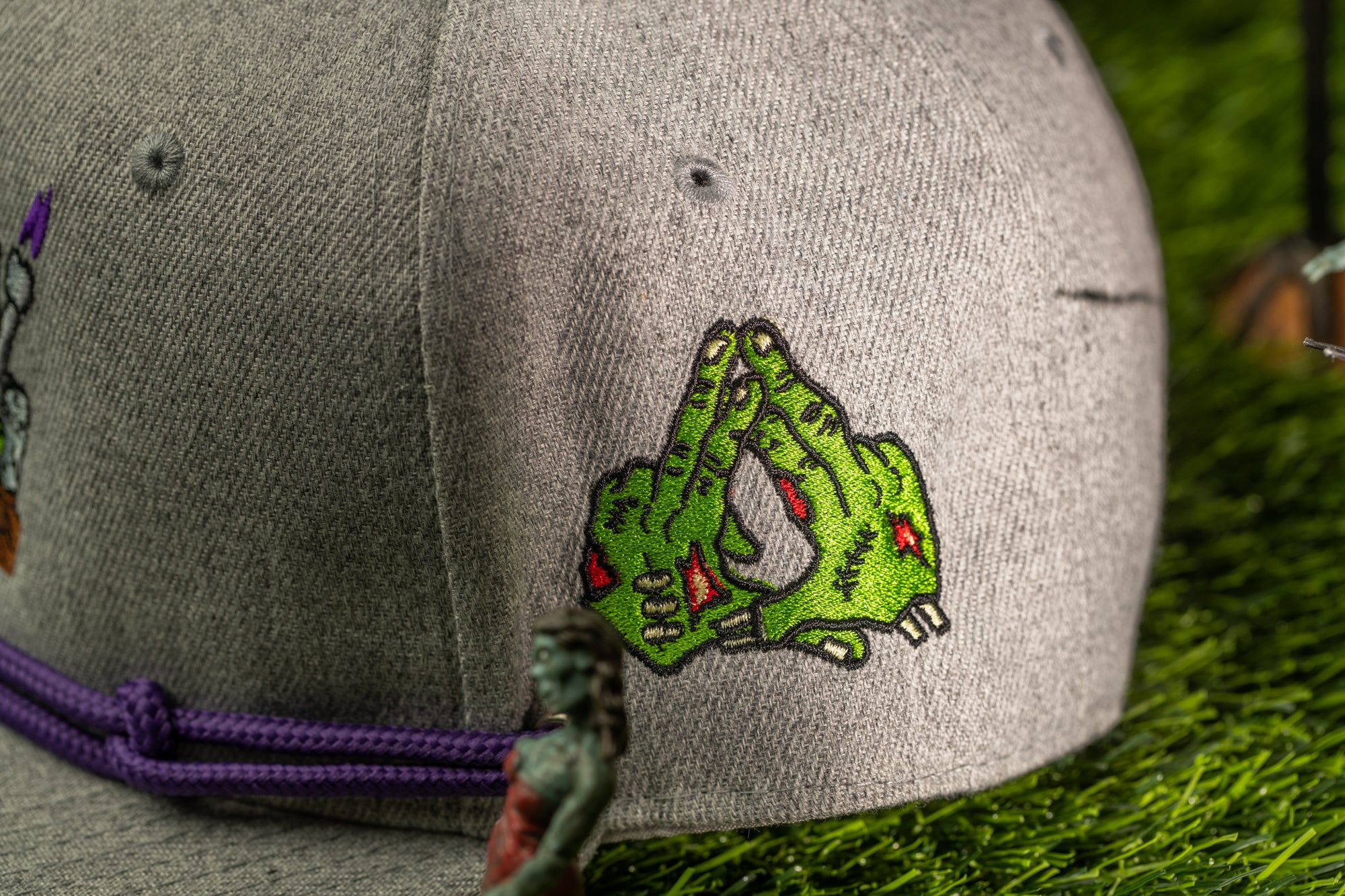 A close-up view of a gray baseball cap with a green zombie hand embroidered on it, placed on a grassy surface.
