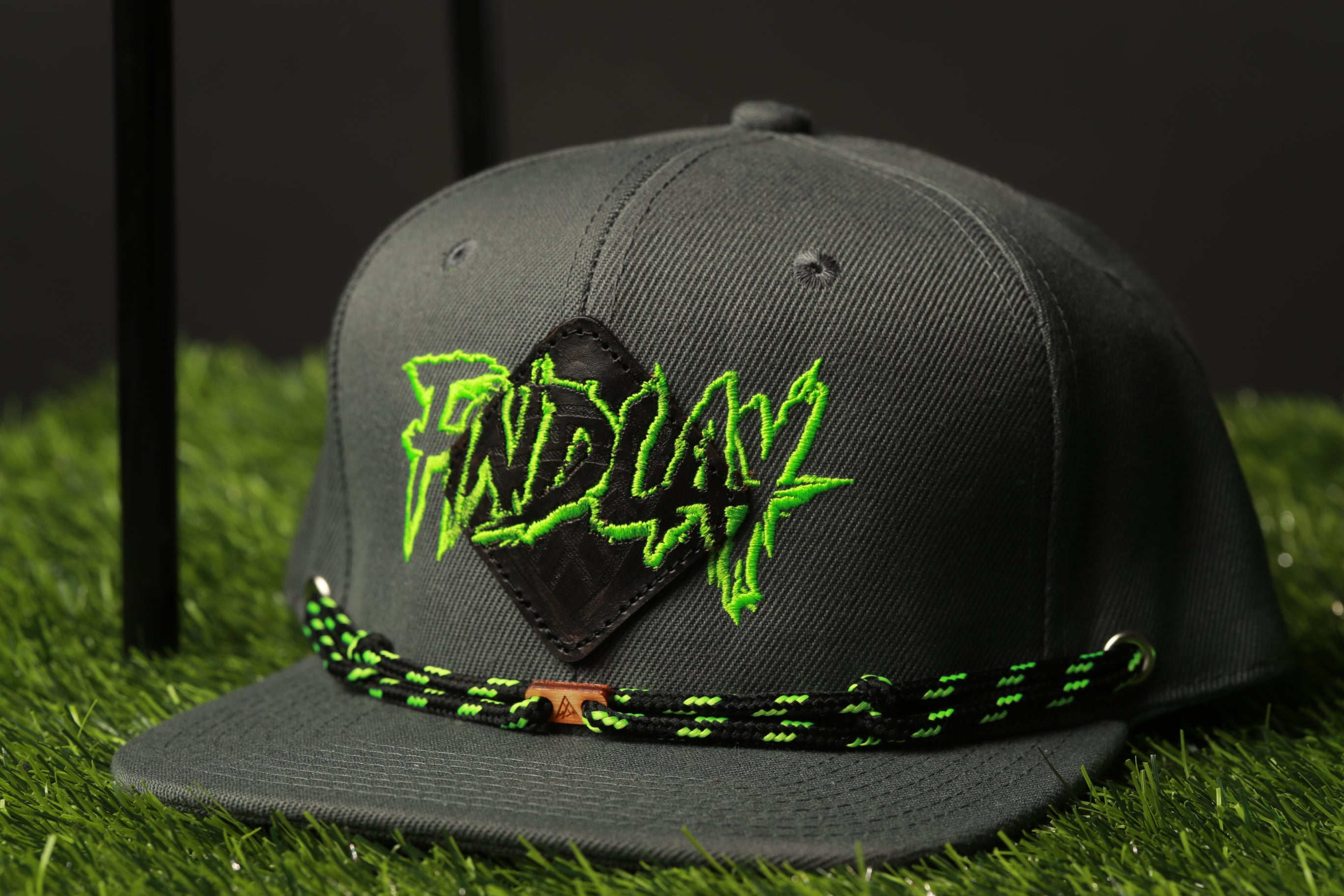 A black baseball cap with a green and black braided strap, featuring a large green and black logo on the front.