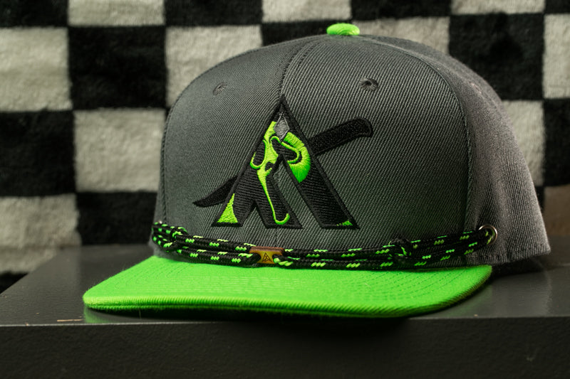 A grey baseball cap with a green brim and a black and green logo on the front, displayed on a black surface against a checkered background.