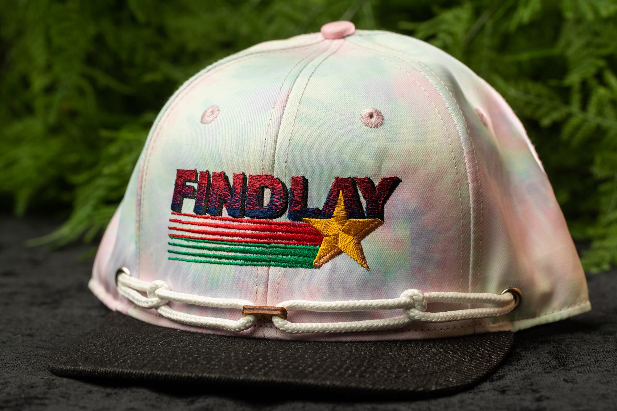 A tie-dye baseball cap with the word "FINDlay" printed on it, set against a blurred green background.